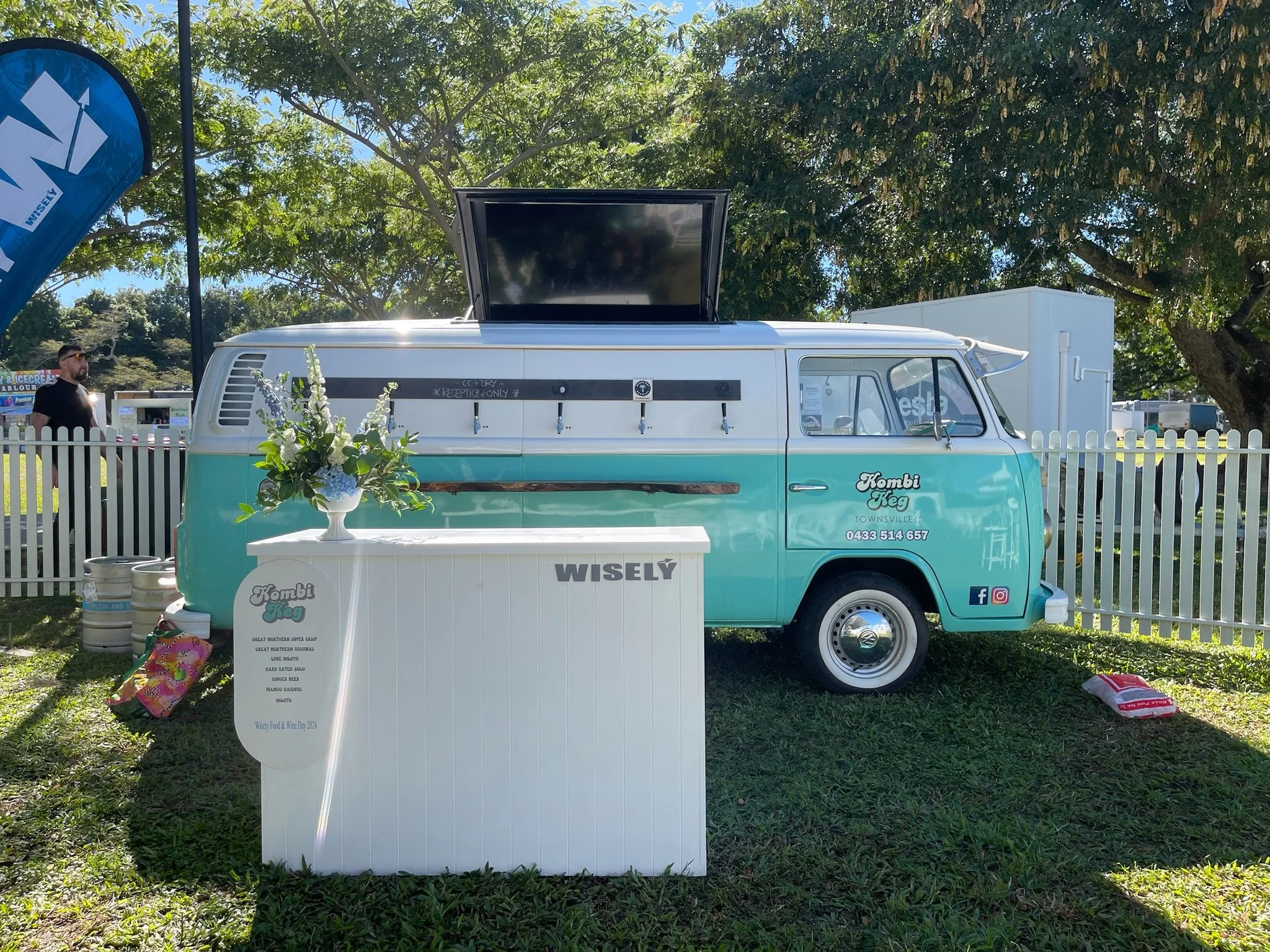 A turquoise and white vintage ice cream truck, called 'Kombi Keg,' parked on grass next to a white fence at an outdoor festival. The truck has a canopy on top, an open window, and a small white table with a floral arrangement nearby. There are beer k