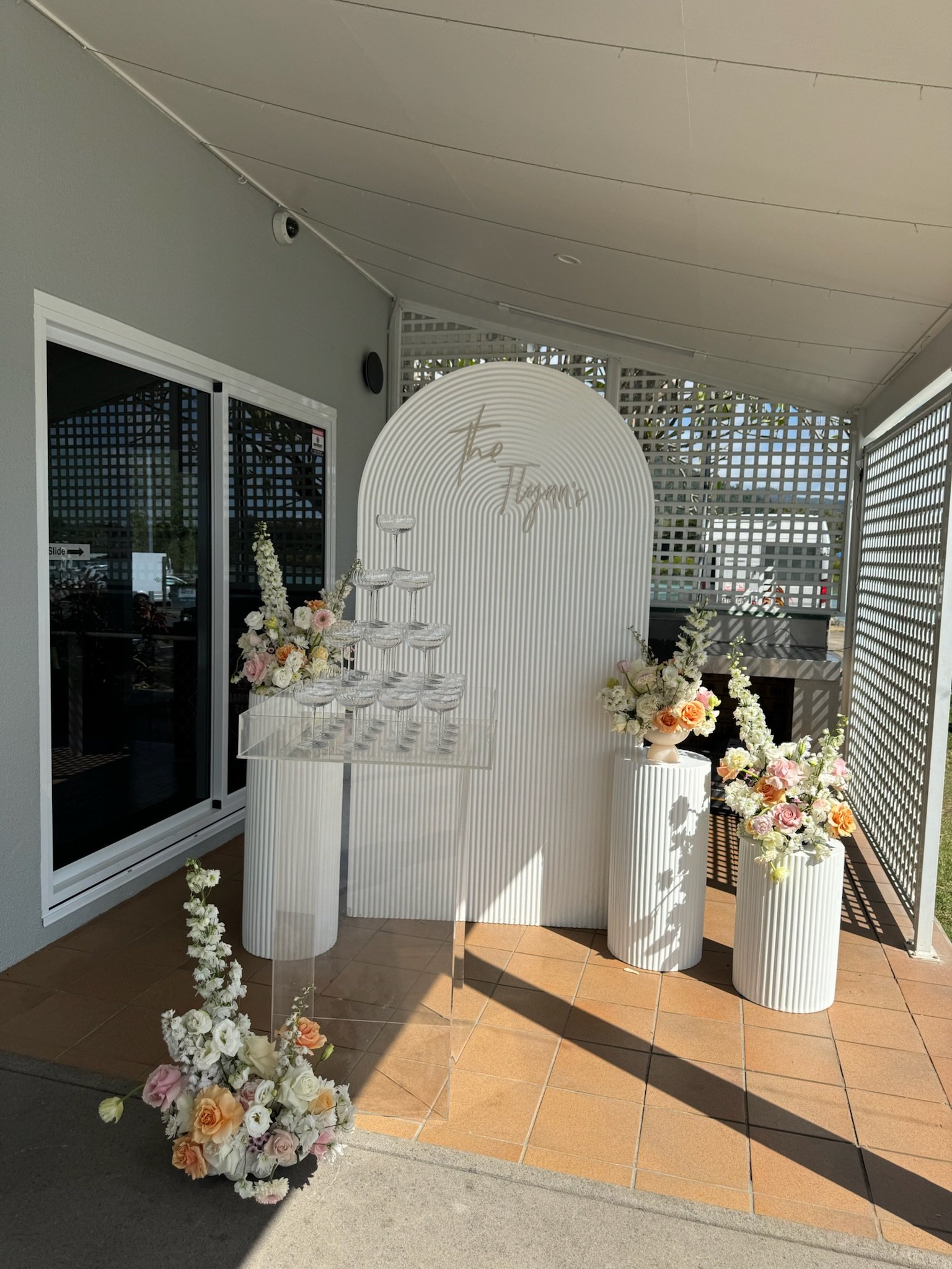 Decorative floral arrangement and champagne glasses on display at an outdoor celebration venue with a white textured backdrop and the words 'The Tapestry' in gold lettering.