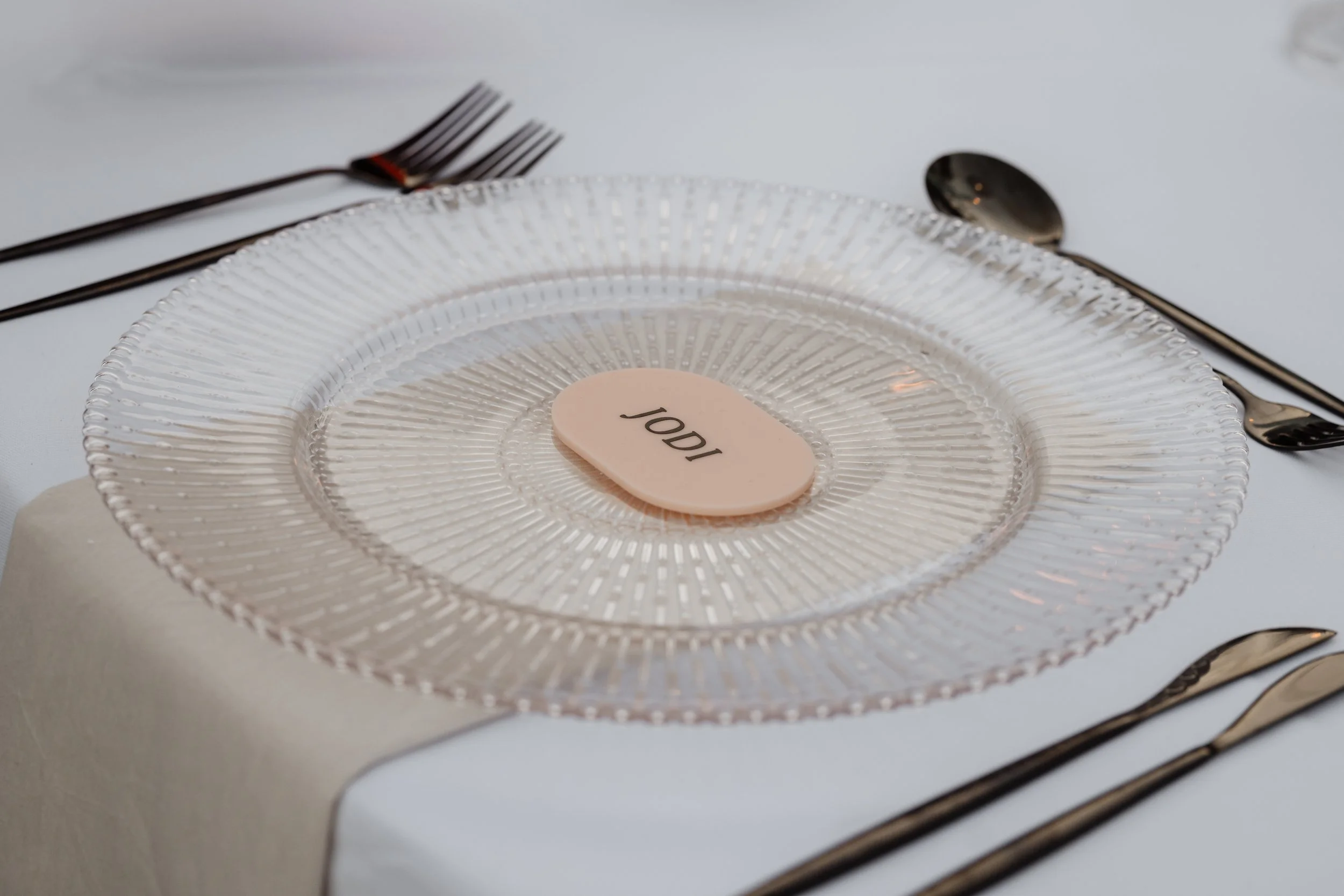 Empty clear decorative glass plate with a pink place card labeled 'JOD' on a white tablecloth, accompanied by silverware.
