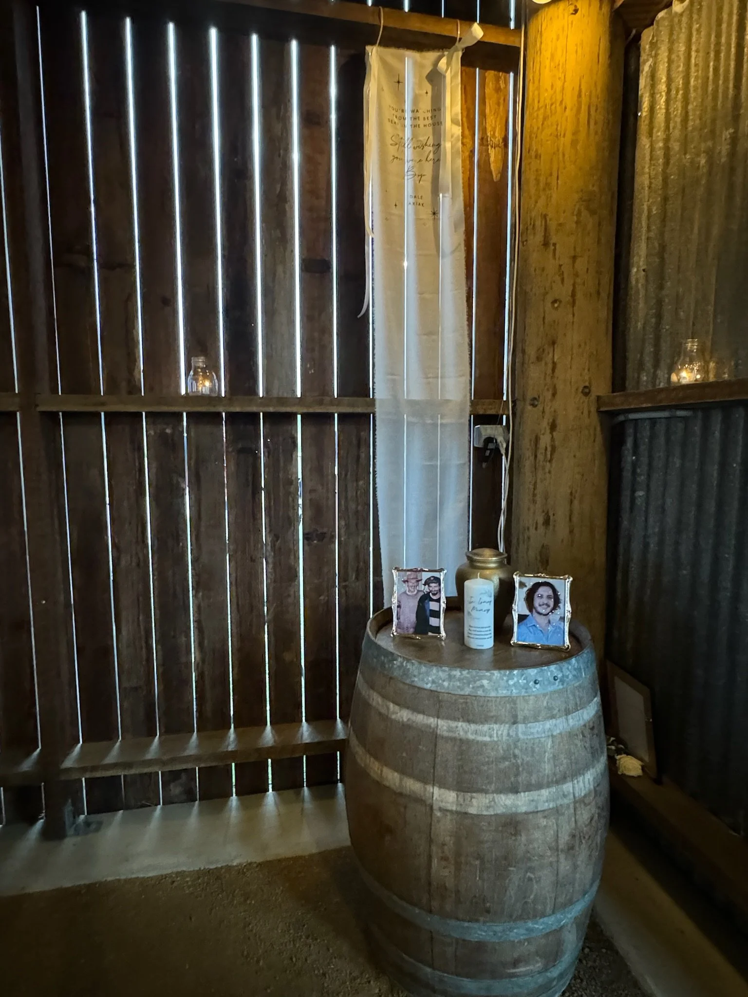 A rustic interior with a wooden barrel used as a table, holding framed photos, a candle, and a jar, set against wooden and metal walls with a white hanging cloth or curtain.