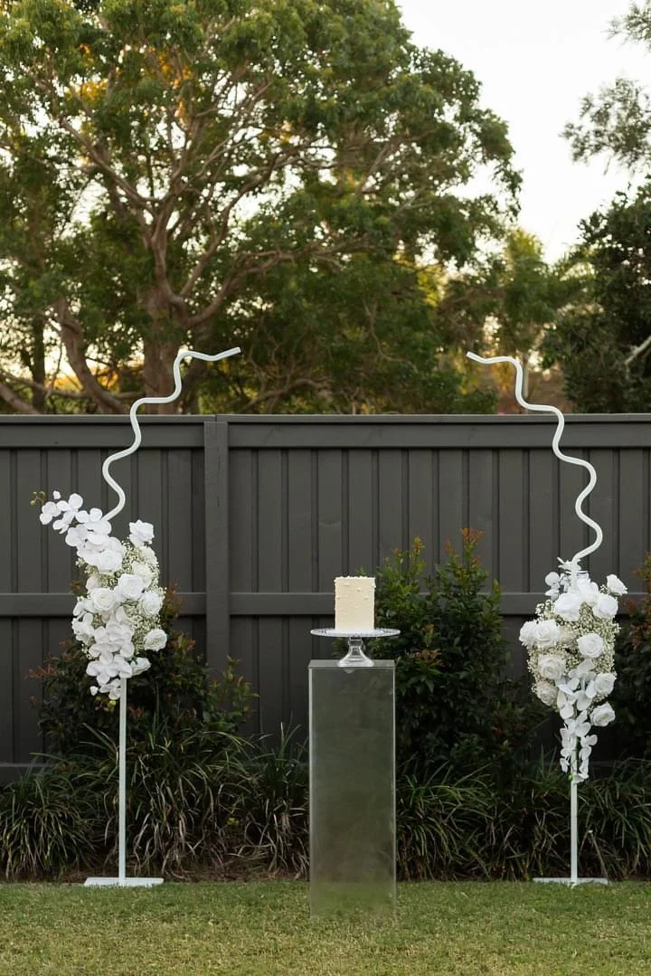 Outdoor wedding or event setup with two tall white floral arrangements, a small white cake on a stand, and a black wood fence in the background.