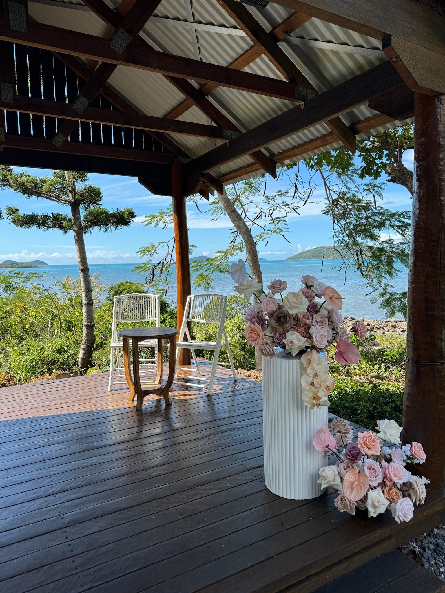 A shaded wooden deck overlooking a body of water with trees and hills in the distance. There are two white chairs, a small round table, and a tall white vase filled with pink, white, and purple flowers on the deck.