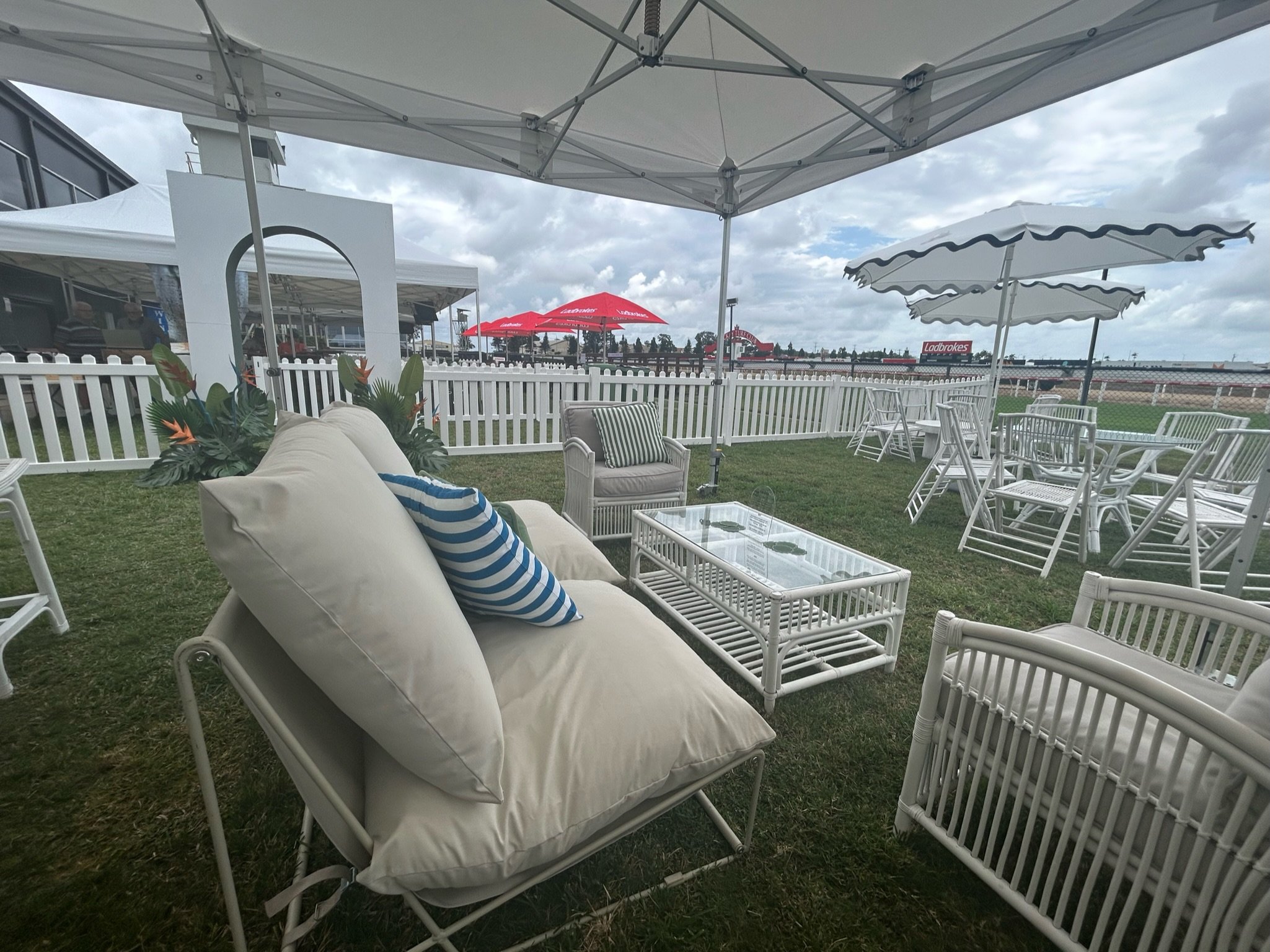 Outdoor seating area with white furniture, umbrellas, and a grassy lawn, near a white fence and modern buildings under cloudy sky.