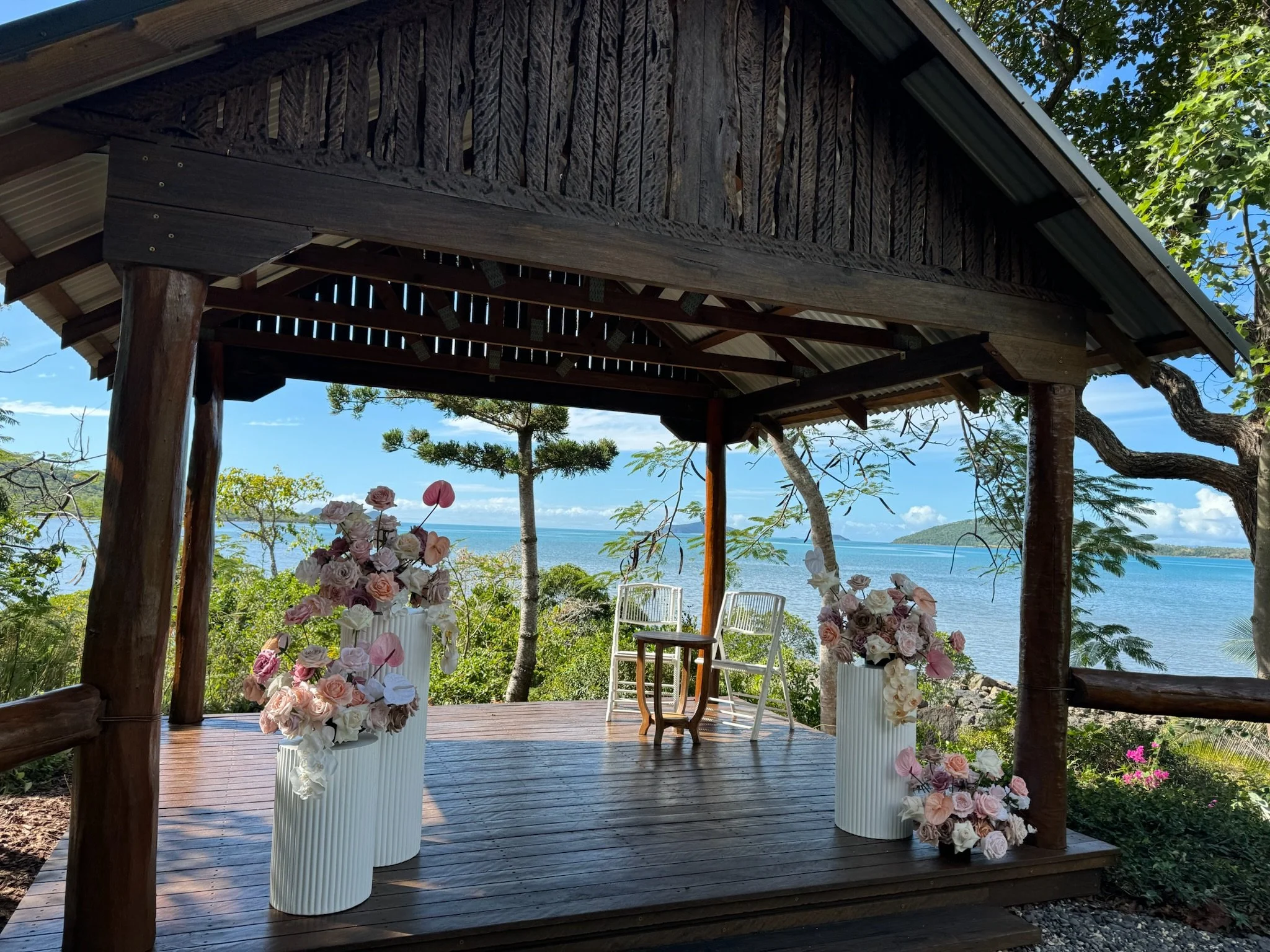 A small wooden gazebo with two white chairs and a small table set up outdoors near the ocean, decorated with pastel-colored flowers in tall white vases.