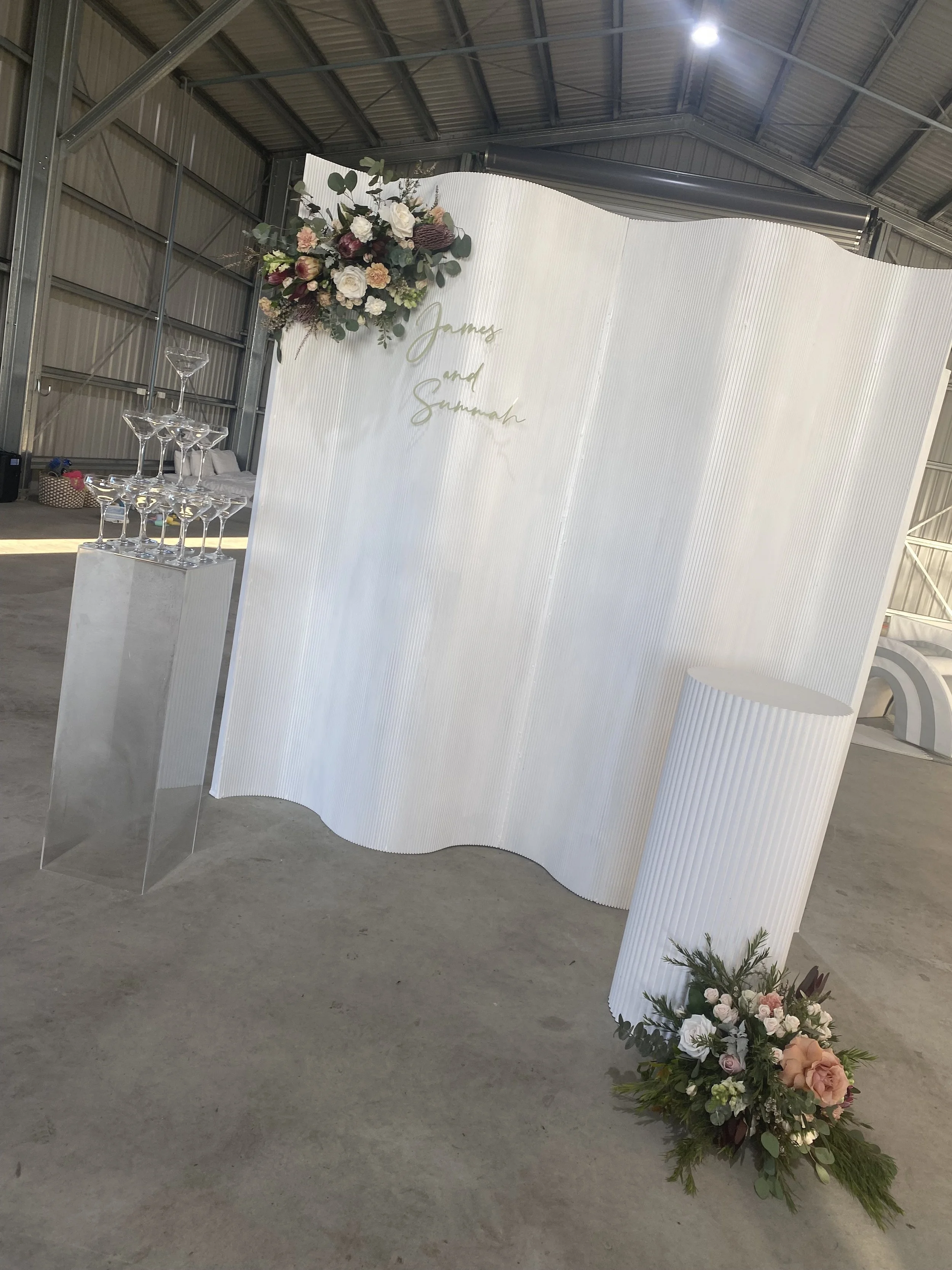 Wedding decoration with a white curved backdrop featuring floral arrangements at the top left and bottom right corners, and a sign with the names 'James and Sumnah' written in gold. There is a clear acrylic stand with a pyramid of champagne glasses o