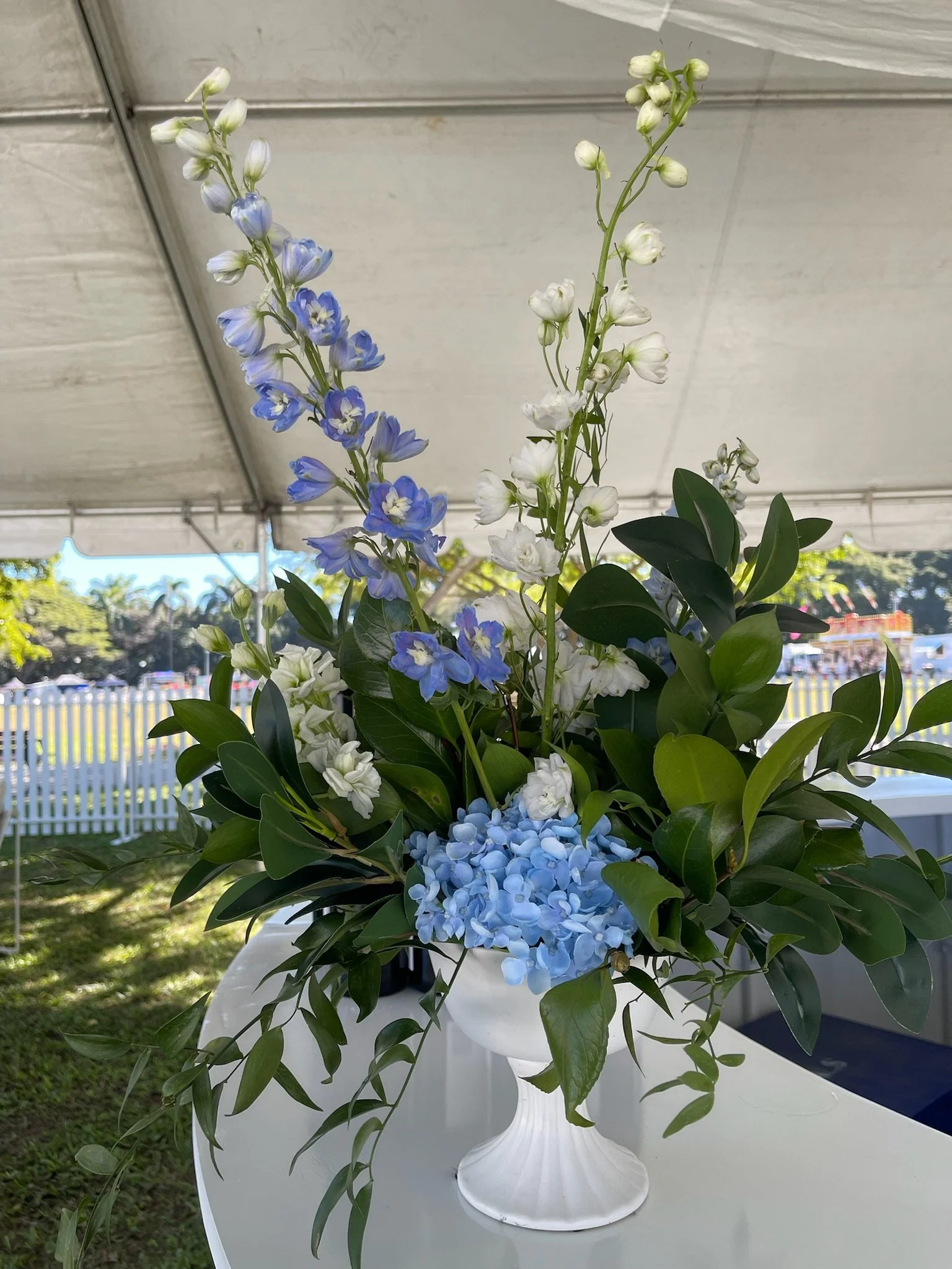 A floral arrangement with white, blue, and purple flowers and green leaves in a white vase, set on a white table under a tent.