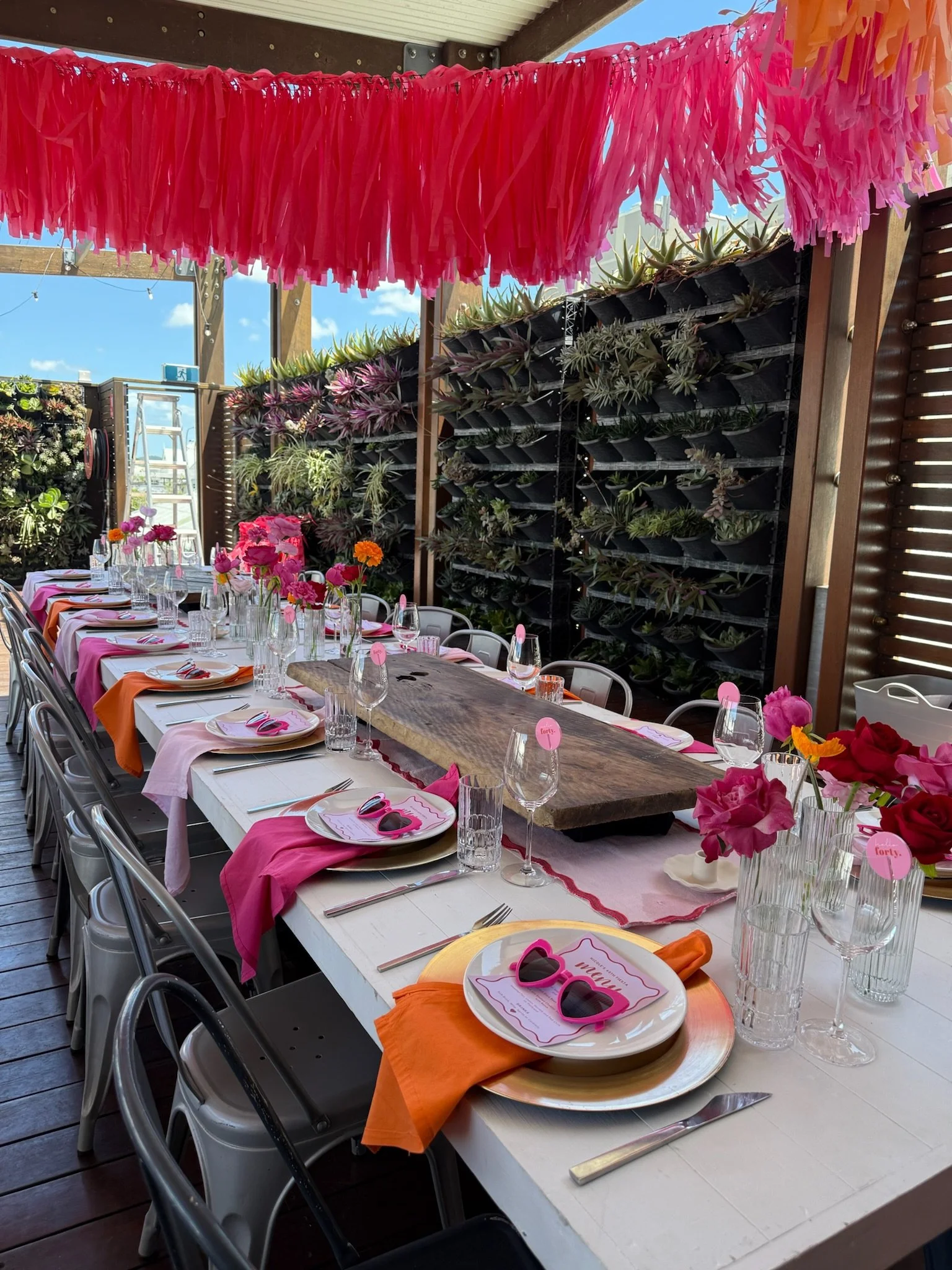 Partially set outdoor party or dining table with pink and orange napkins, glasses, plates with pink sunglasses and pink paper place cards, pink and orange flowers, and pink hanging decorations overhead. There is a vertical garden with succulents in t