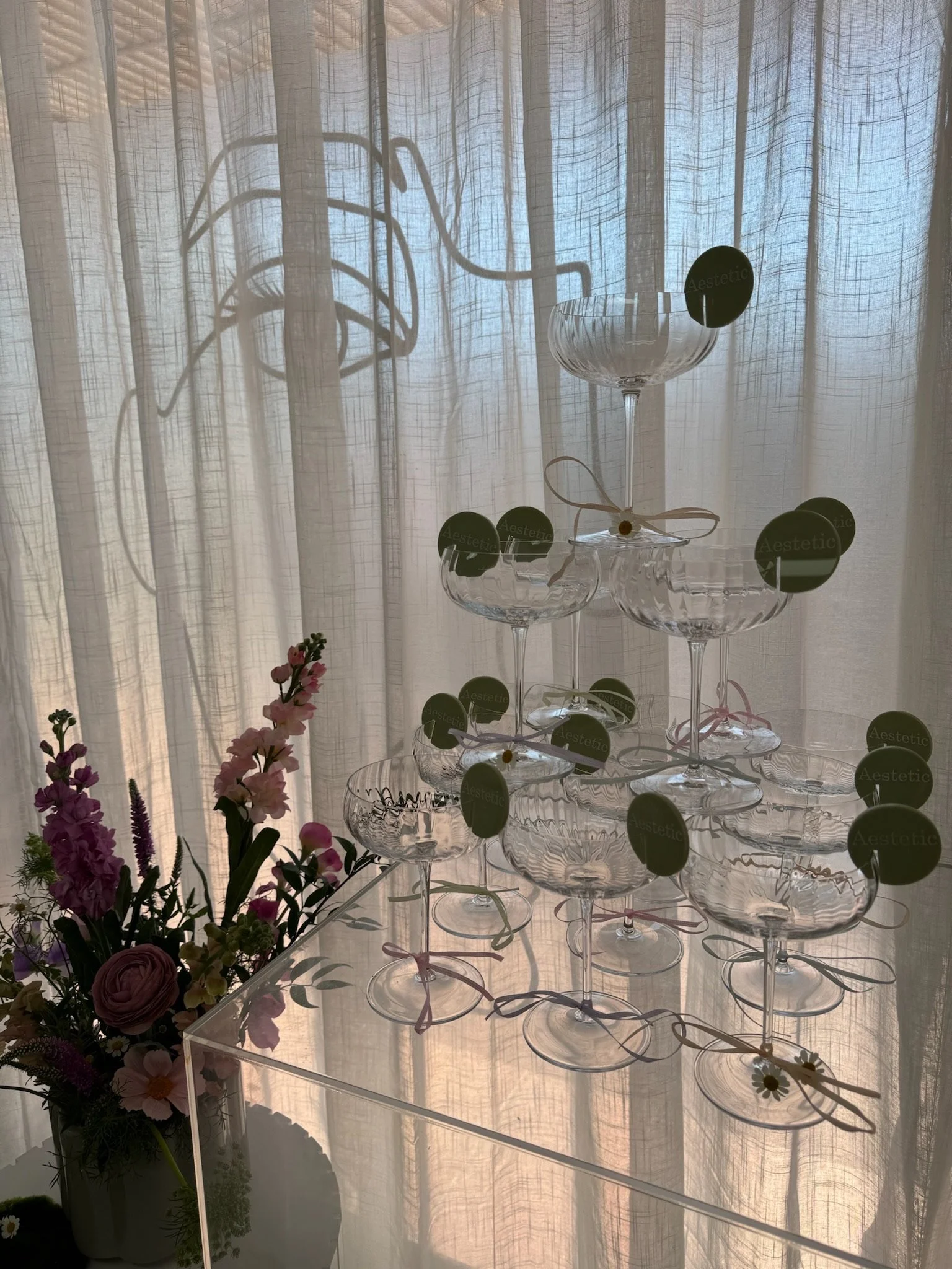 Stacked glass dessert cups with green round tags on a transparent display stand, with a flower arrangement nearby, backlit by soft light through a sheer curtain.