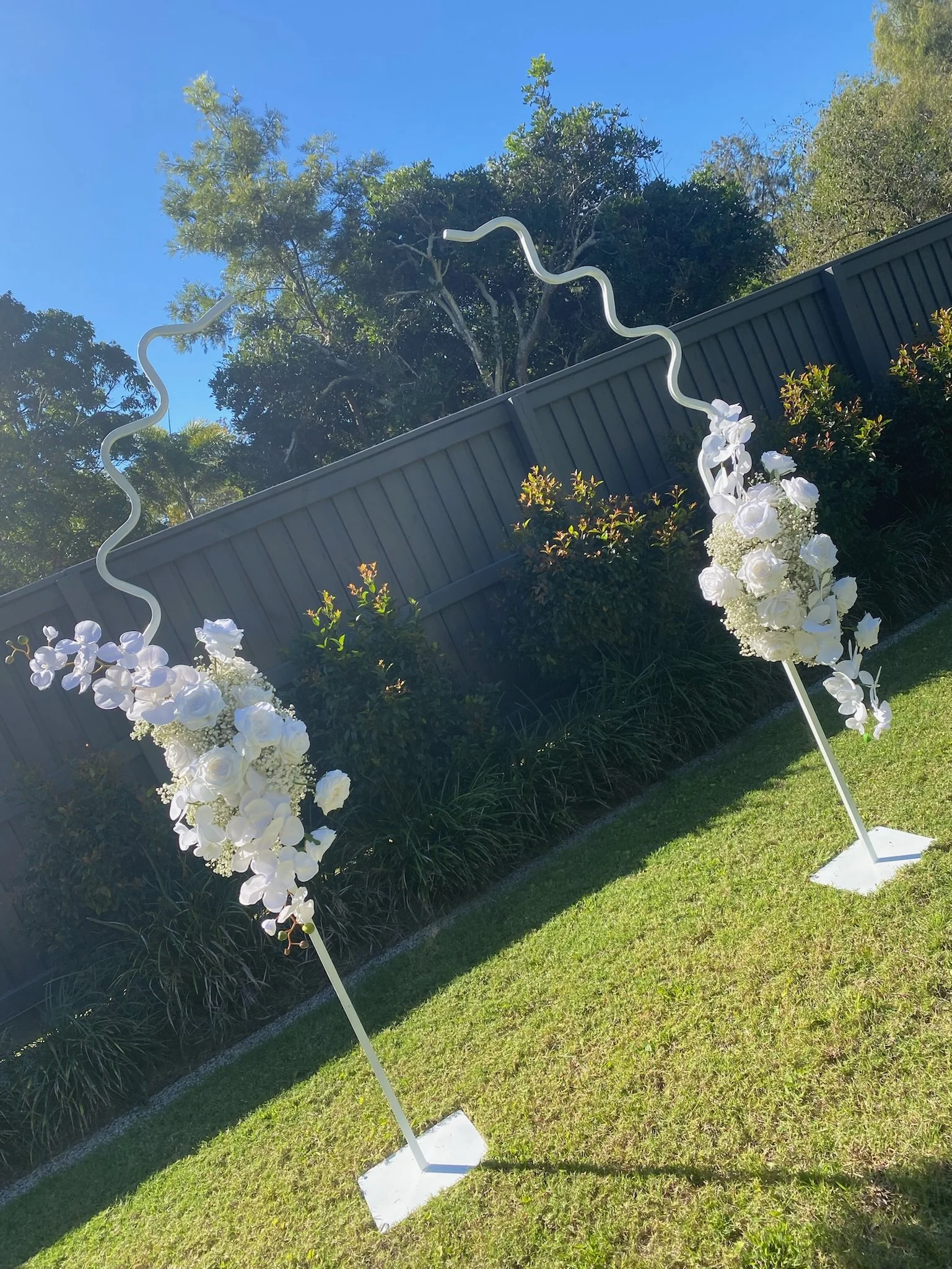 White floral arch with wavy metal sticks on either side, set outdoors on a grassy area with bushes and trees in the background.