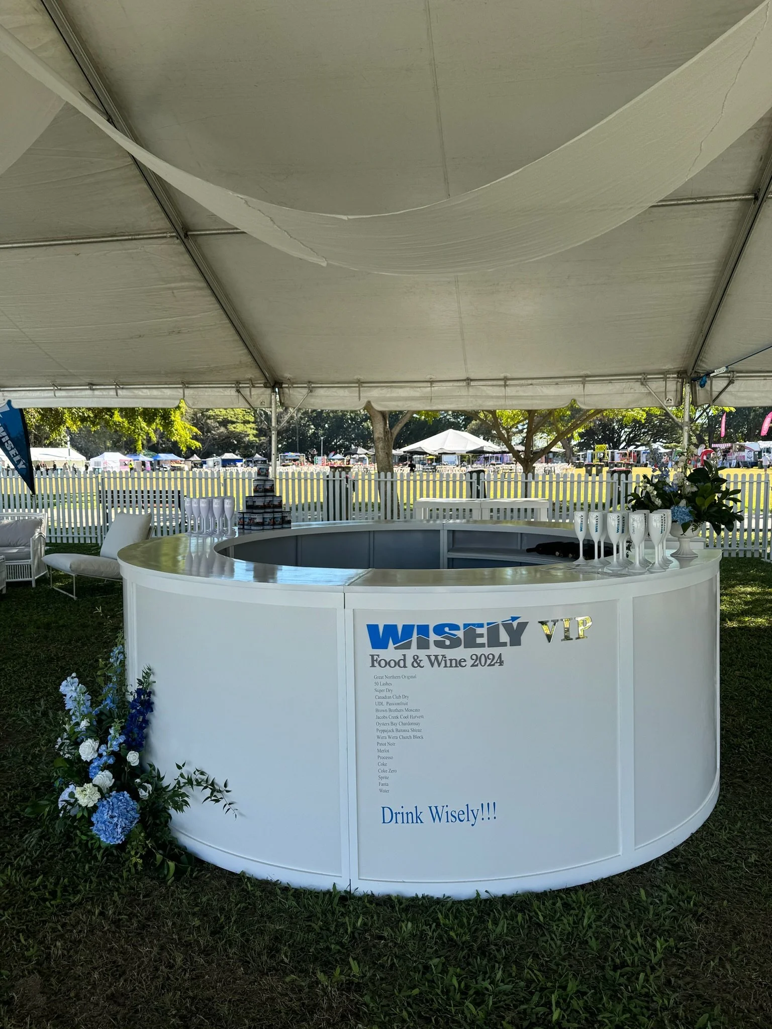 A white circular VIP bar under a tent at Wisely Food & Wine 2024 event, with wine glasses, flowers, and a list of beverages, set outdoors on grass with trees and tents in the background.
