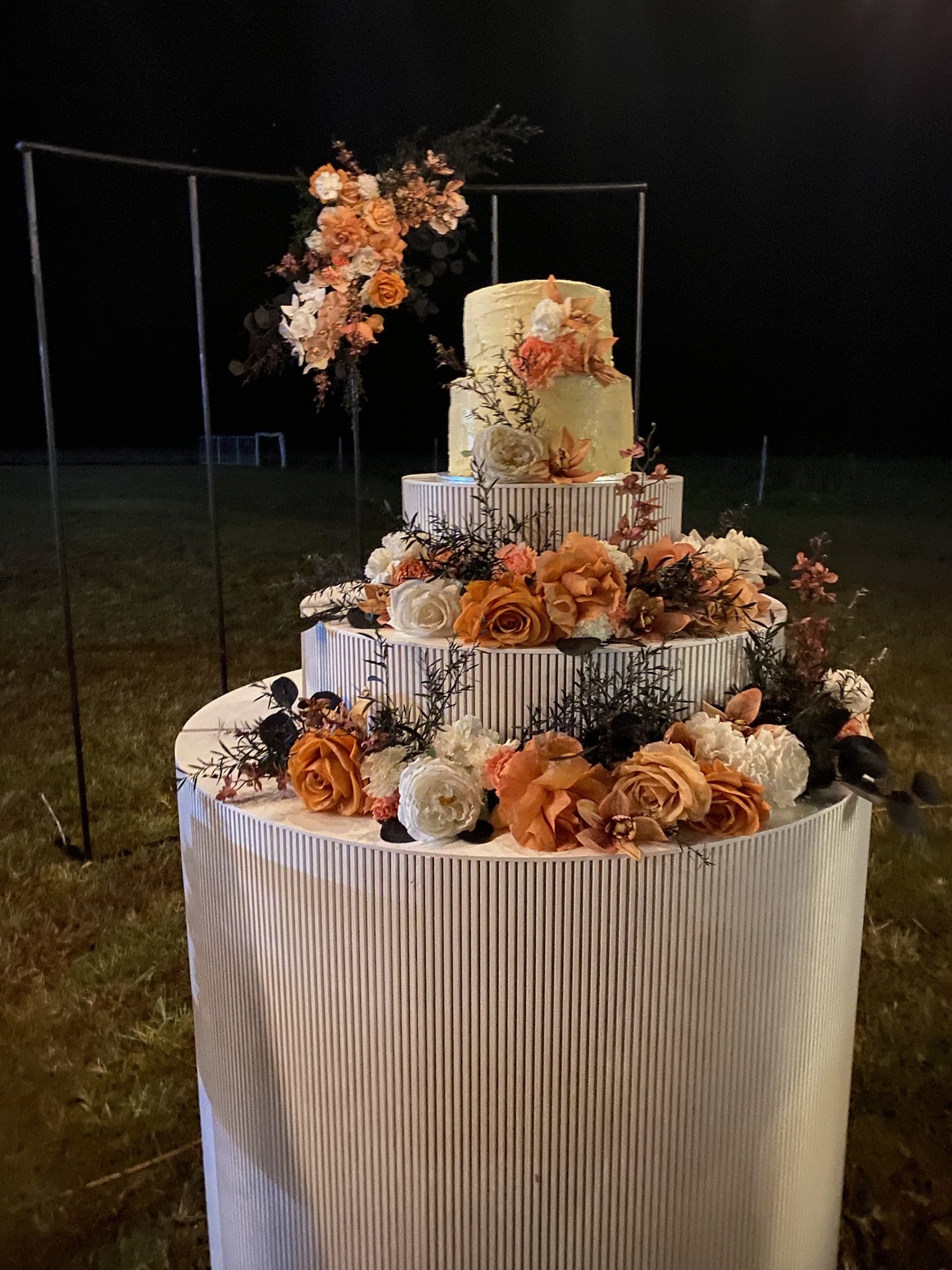 A tiered wedding cake decorated with white and peach-colored flowers and greenery, set outdoors at night with a dark sky background.