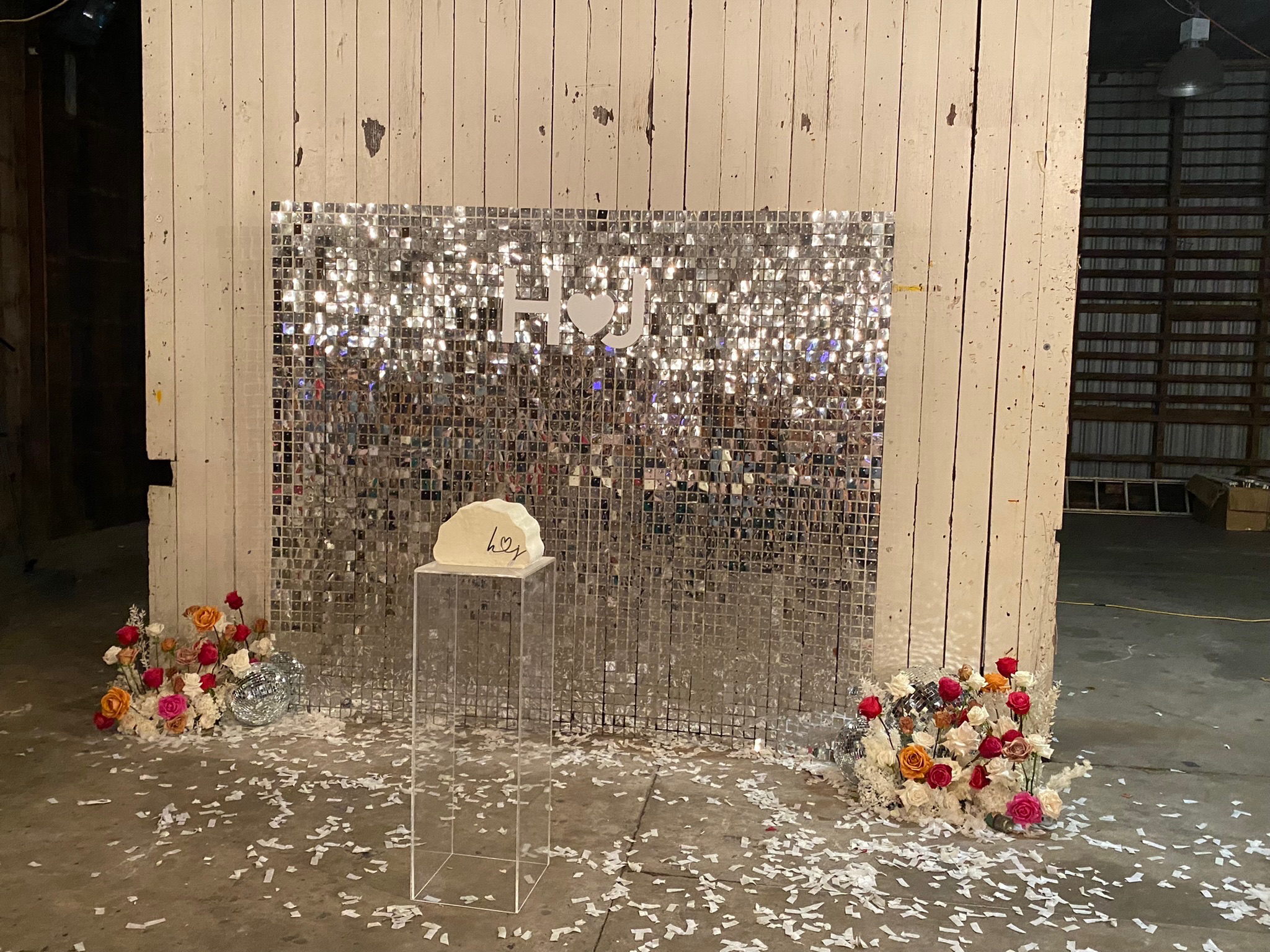 Decorative setup with a sparkly mirrored backdrop, floral arrangements, and a sign that says 'Hey' in an industrial space.