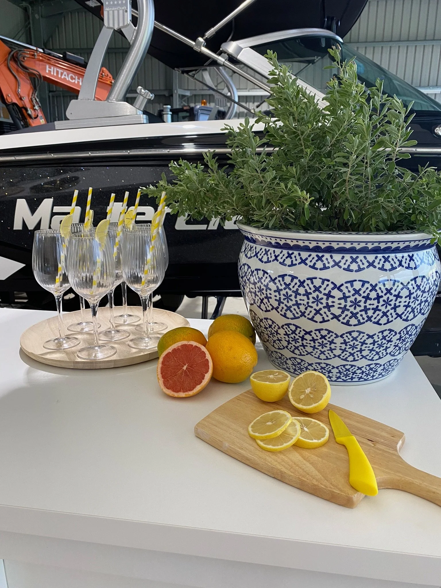 A table setup with four empty champagne glasses with yellow striped straws on a round wooden tray, a cutting board with sliced lemons and a yellow knife, a large blue and white decorative pot with green herb, and a bunch of fresh citrus fruits includ