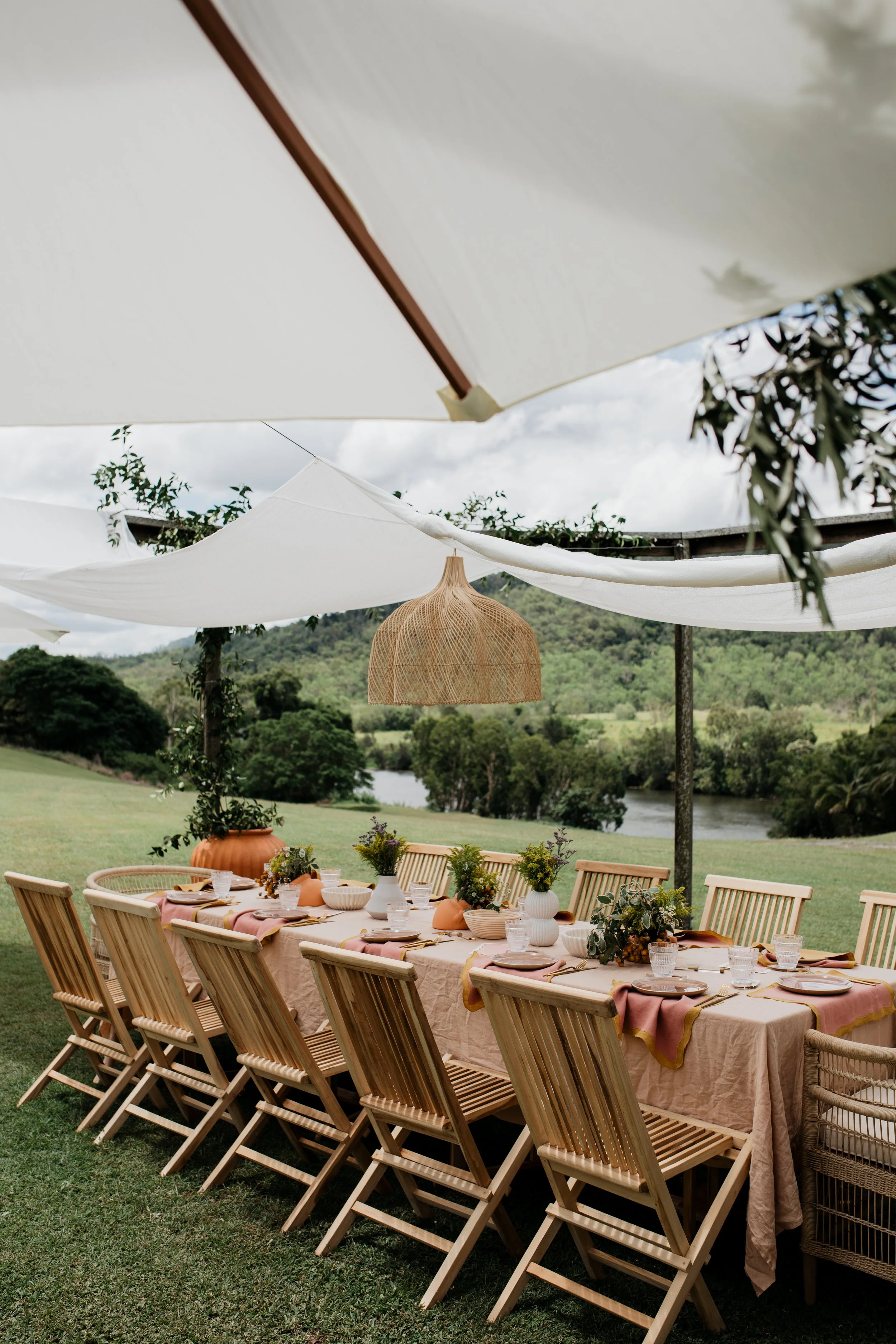 Outdoor dining table set for a meal, decorated with vases and flowers, situated on a lush green lawn with a river and trees in the background, shaded by large white umbrellas and a woven hanging light.