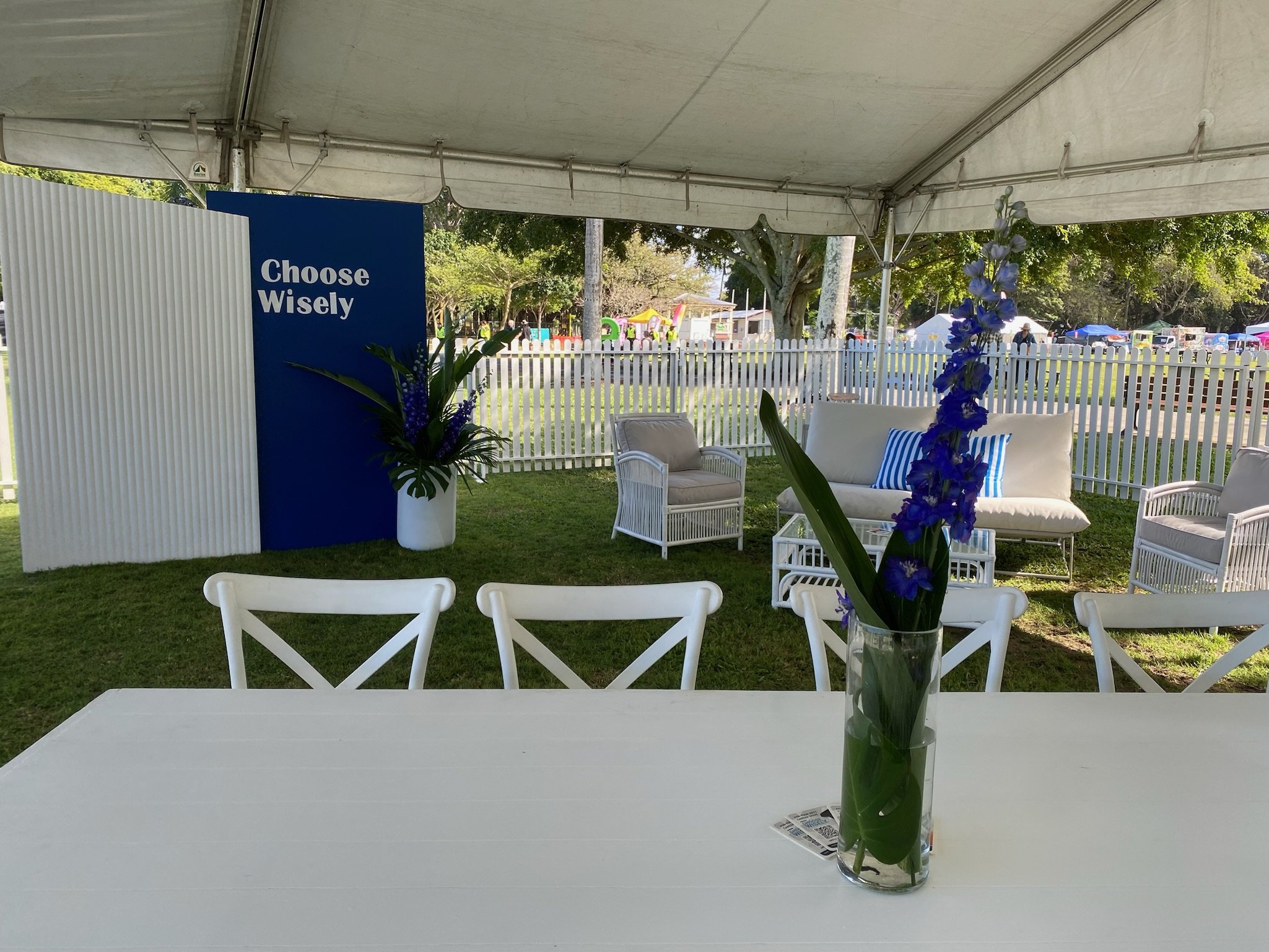 Indoor tent with white tables and chairs, a blue and white sign reading 'Choose Wisely,' vases with blue flowers, and outdoor park with trees and tents in the background.