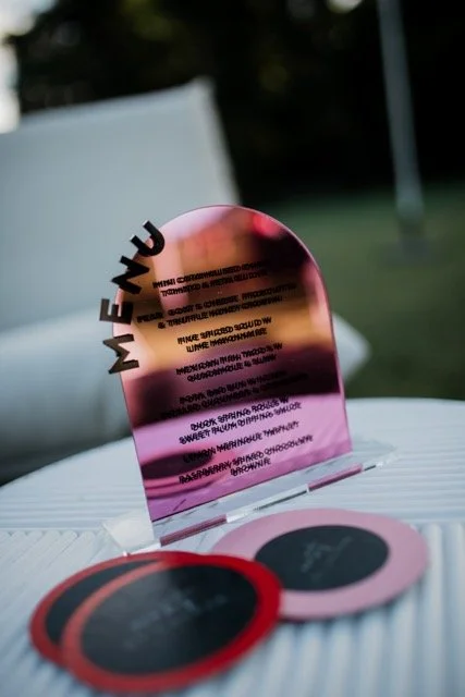 A pink reflective oval sign with black text that reads 'ME + U' and additional blurred text below, placed on a white surface outdoors.