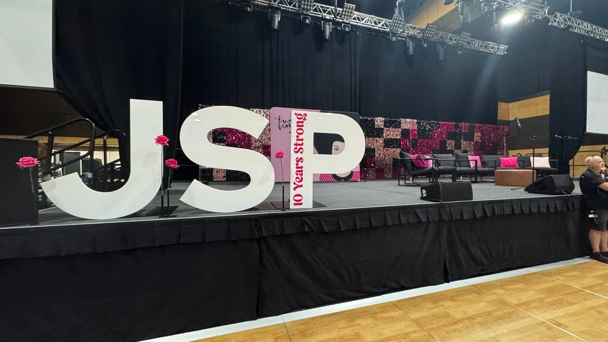 Empty stage decorated with large white letters 'J', 'S', and 'P' with pink flowers attached. A vertical banner between 'S' and 'P' reads '10 Years Strong' in red text. The stage has black curtains, a patterned backdrop, black chairs with pink pillows