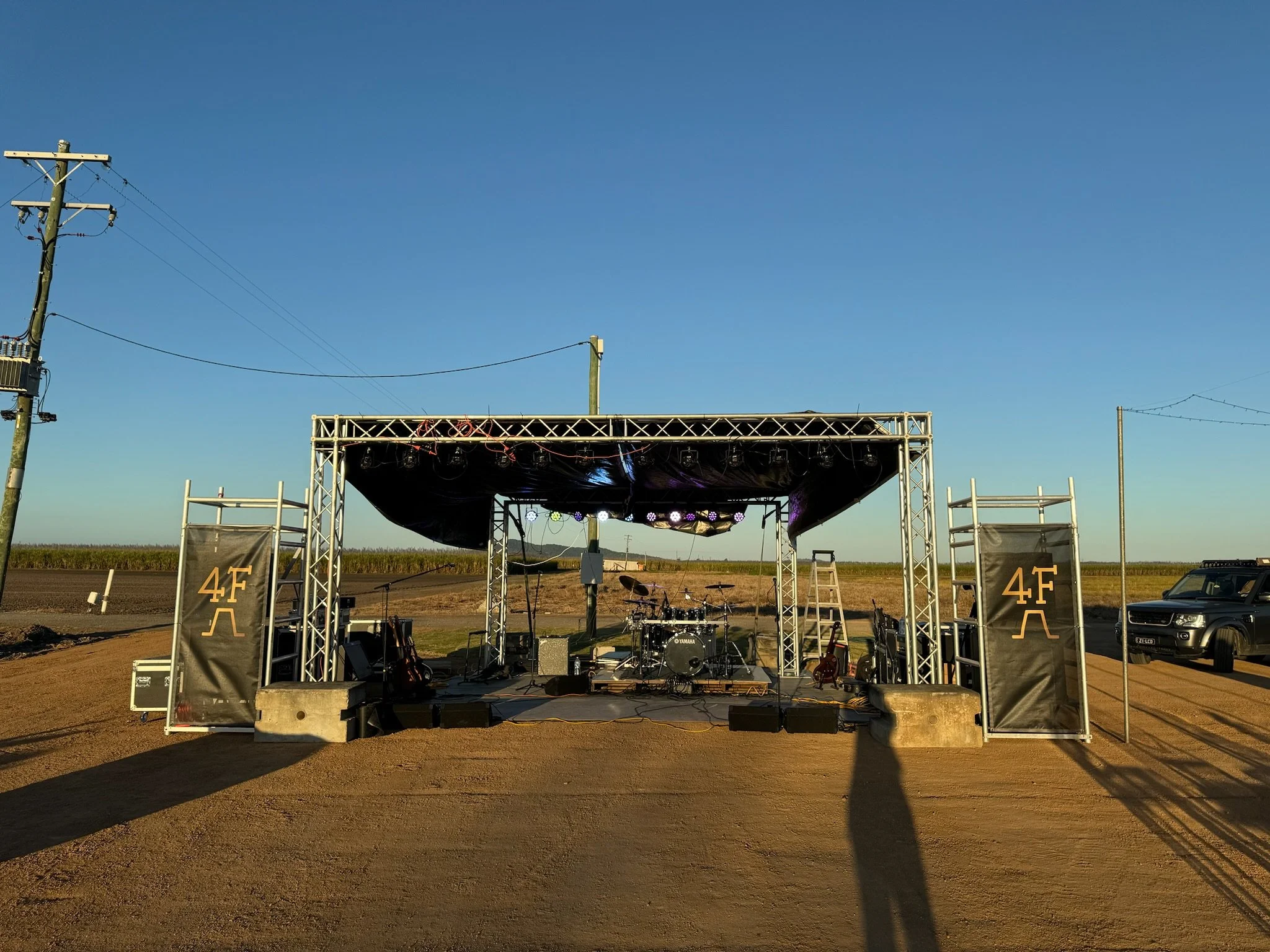 An outdoor music stage set up on a dirt road in a rural area with a clear blue sky, equipped with drums and musical instruments, flanked by black banners with the text "4F" and an abstract logo.
