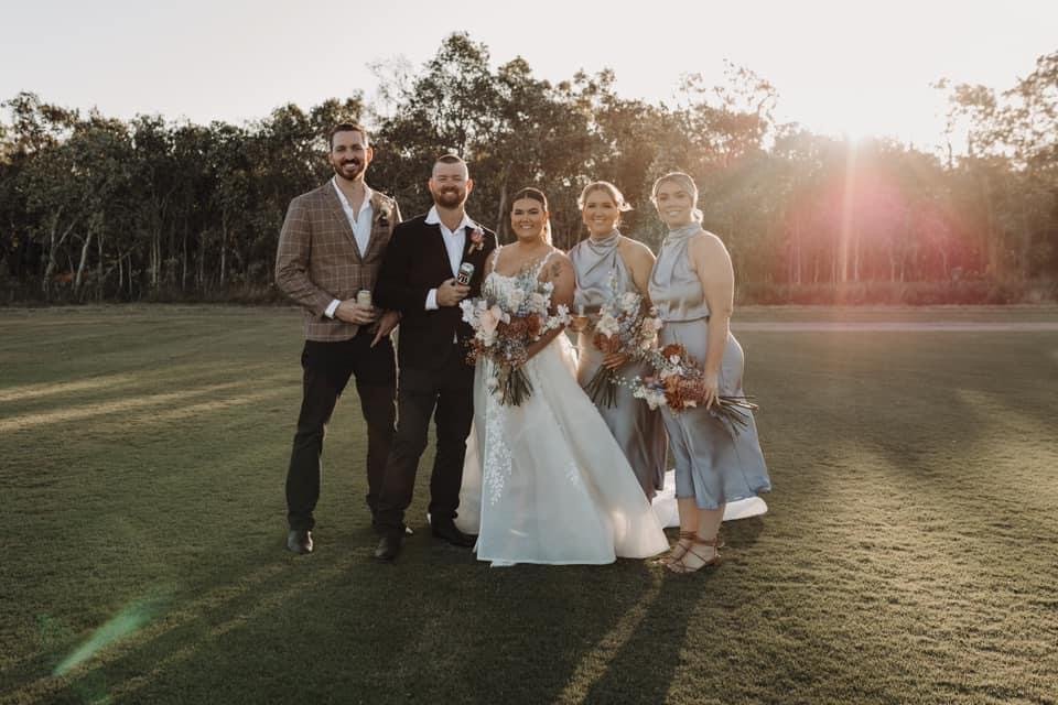 A wedding photo taken outdoors during sunset featuring five people: a bride in a white gown holding a bouquet, two men in suits, and two women in matching dresses also holding bouquets, standing on a grassy field with trees in the background.