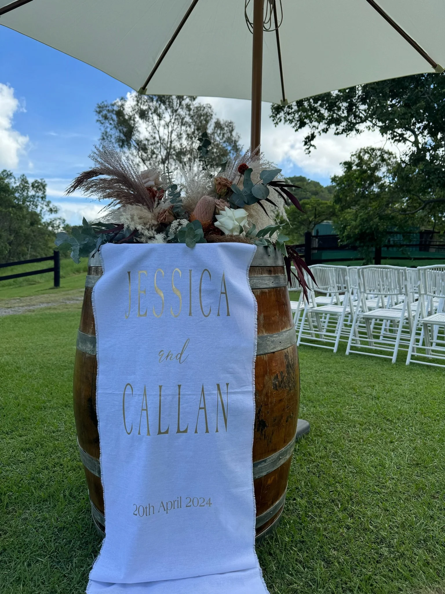 Wedding decor with a wooden barrel covered with flowers and a white cloth that has "Jessica and Callan" written on it, along with the date "20th April 2024". The setup is outdoors with white chairs and a large umbrella, trees, and a clear sky in the 