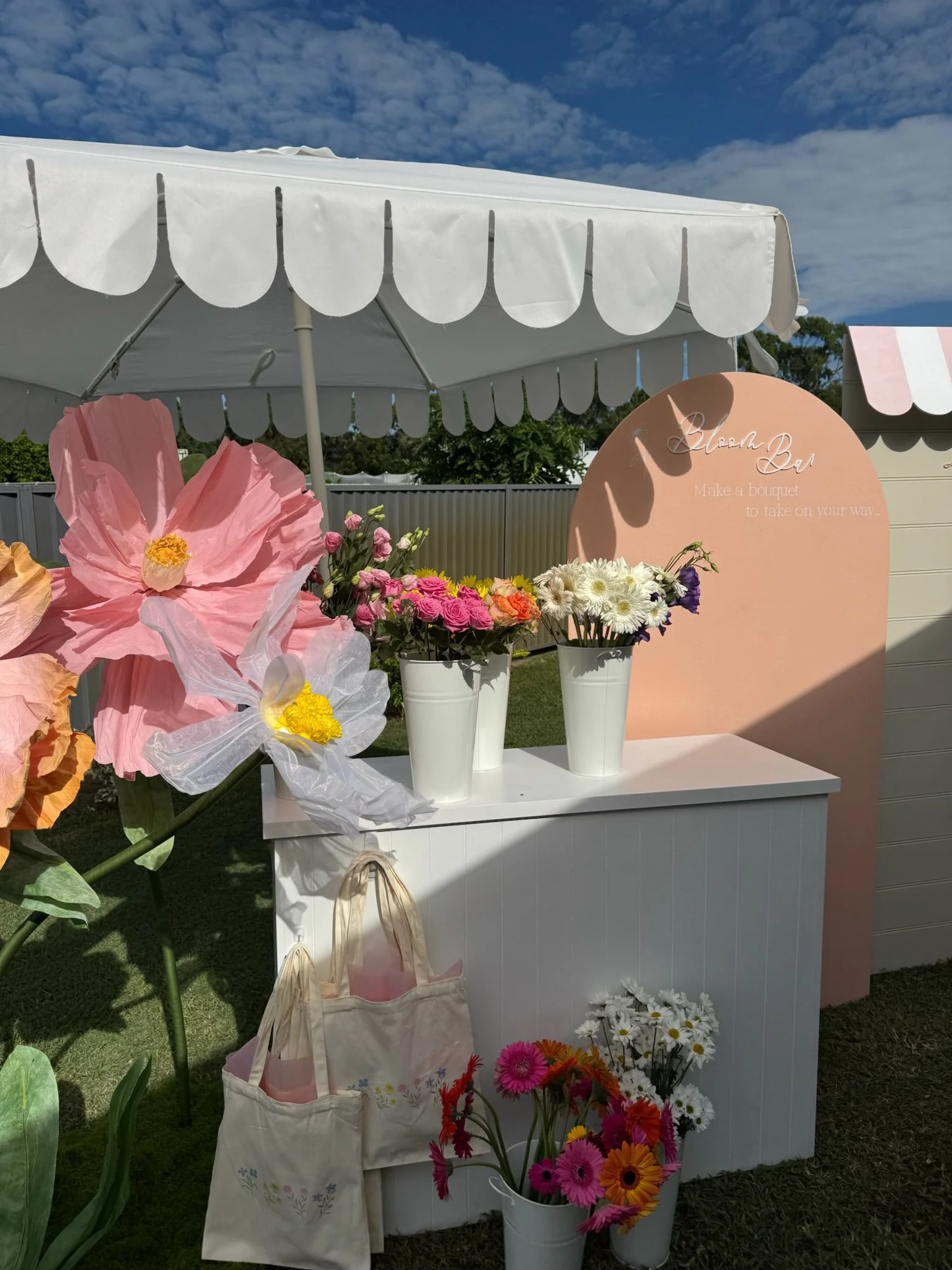 A flower stand at an outdoor event with a variety of colorful flowers in vases, a white cabinet, tote bags, and a sign that reads 'Bloom Bar Make a bouquet to take on your way.' There is a white canopy tent and a blue sky with scattered clouds.