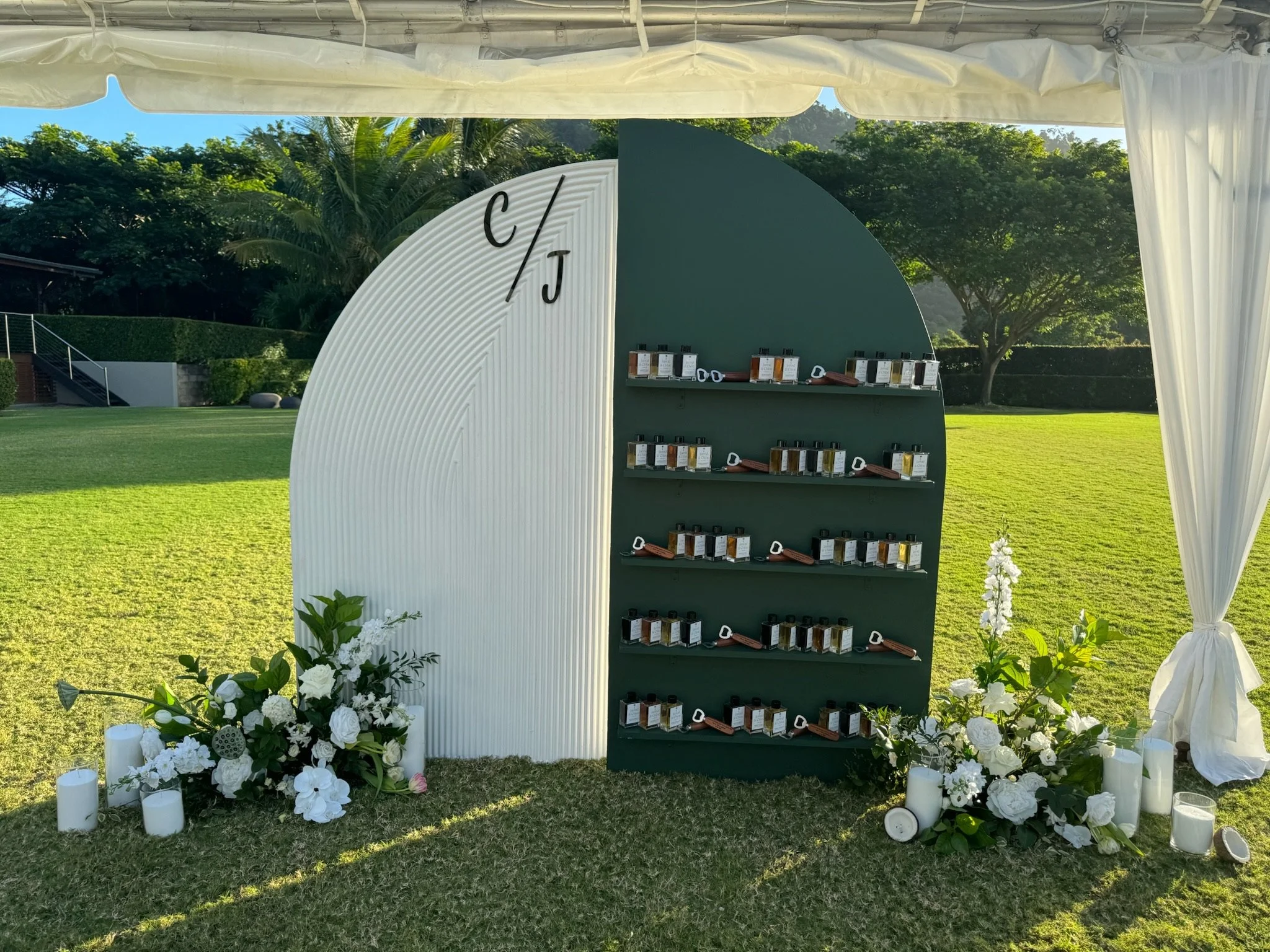 An outdoor display booth with two shelves of perfume bottles and white floral decorations, candles, and greenery on grass with trees in the background.