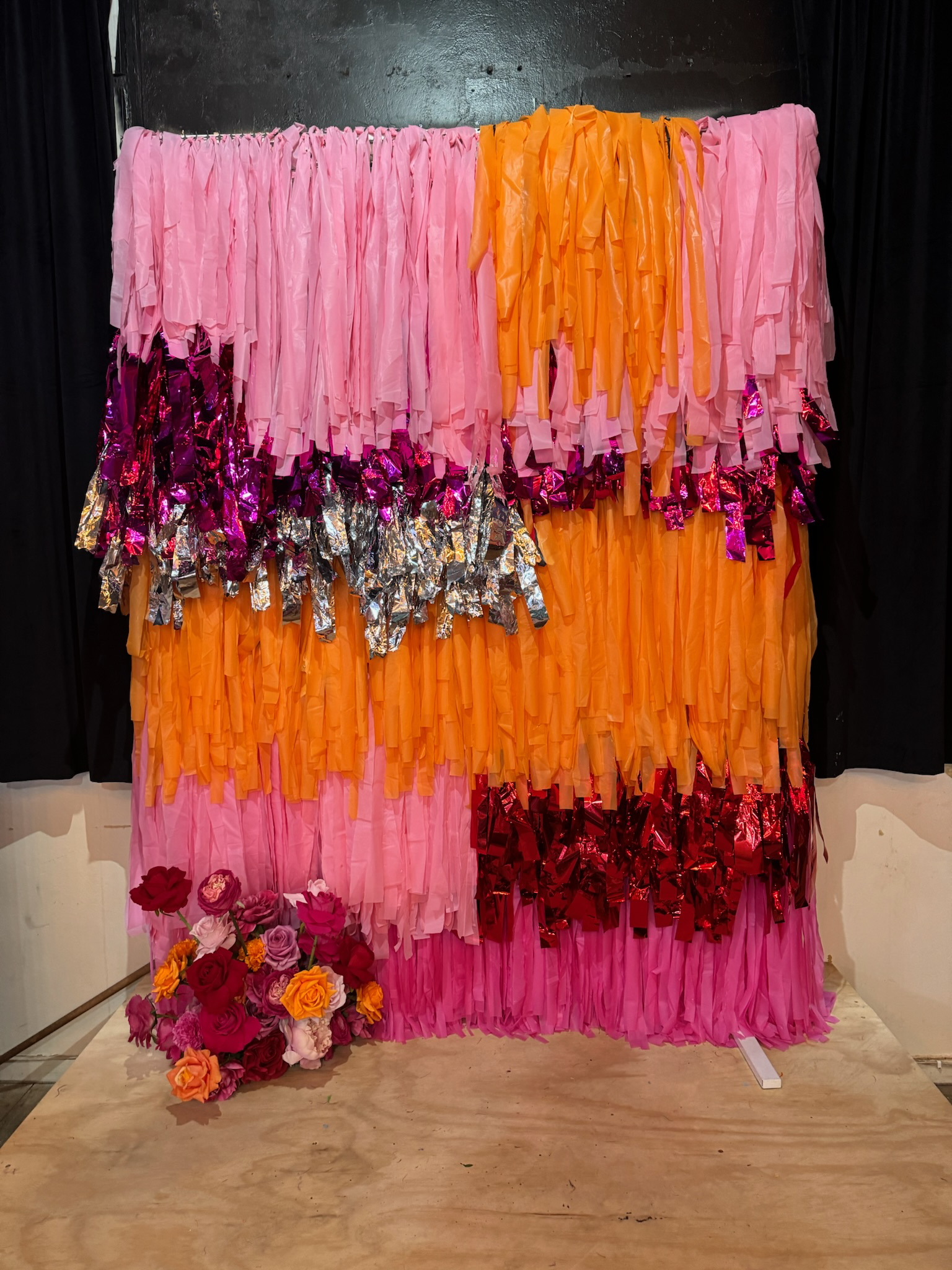 Colorful backdrop made of pink, orange, purple, silver, and red tissue paper strips with a bouquet of artificial flowers at the bottom left.