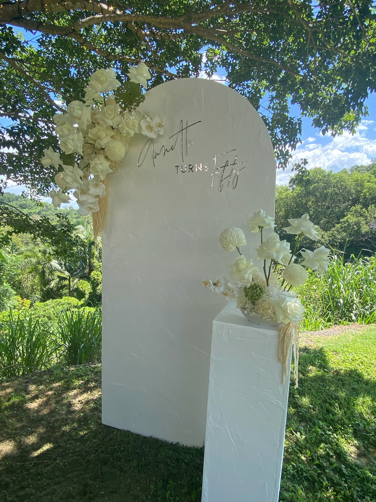 Outdoor wedding or event decoration featuring a white, textured arched backdrop with modern script and serif lettering that reads 'Ariel H. Turns 10.' The backdrop is adorned with white flowers, including orchids and roses, on the upper left corner a