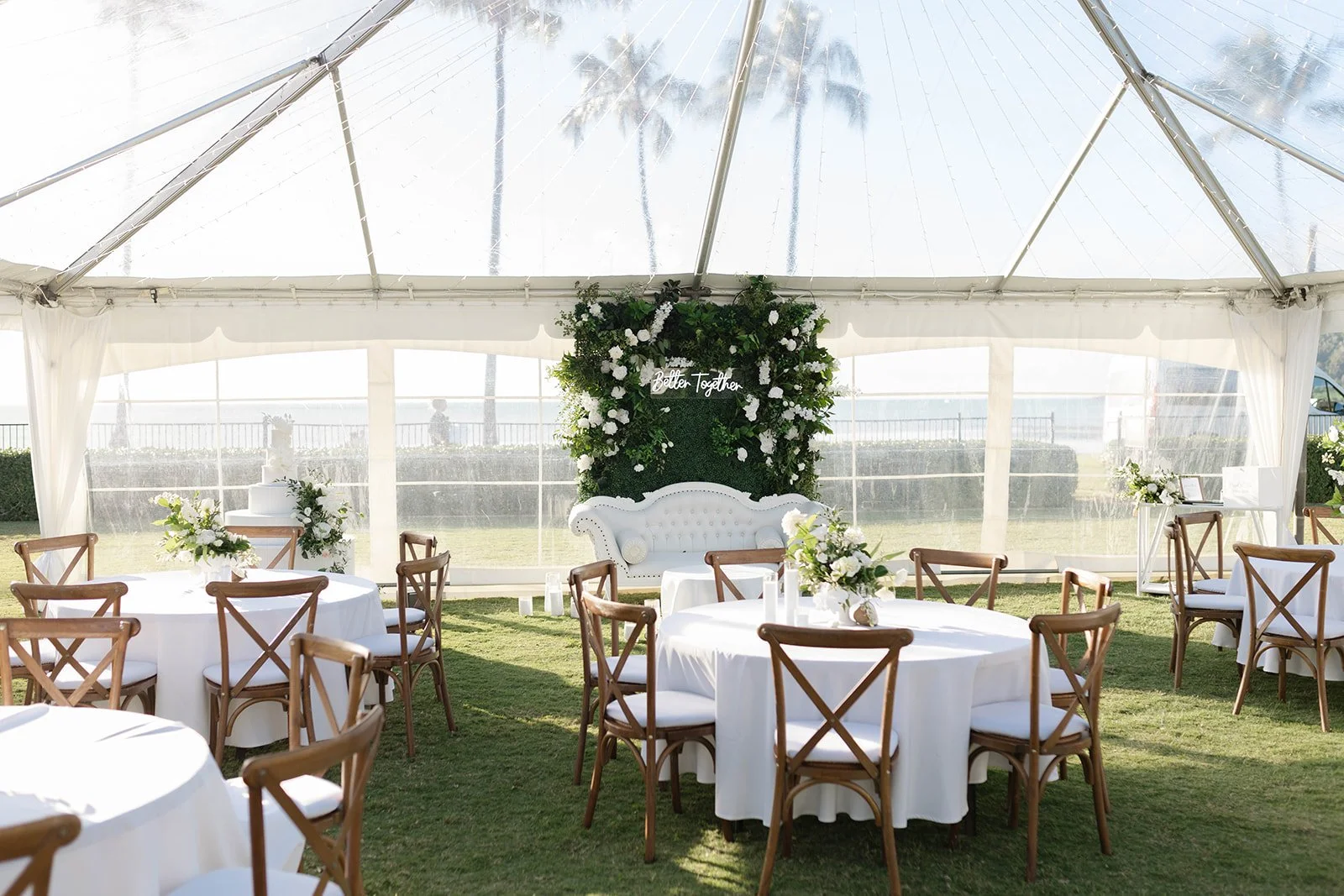 Wedding reception setup inside a white tent with round tables covered in white tablecloths, wooden chairs with white cushions, floral centerpieces, a white sofa, and a backdrop with the words "Better Together" in lights.