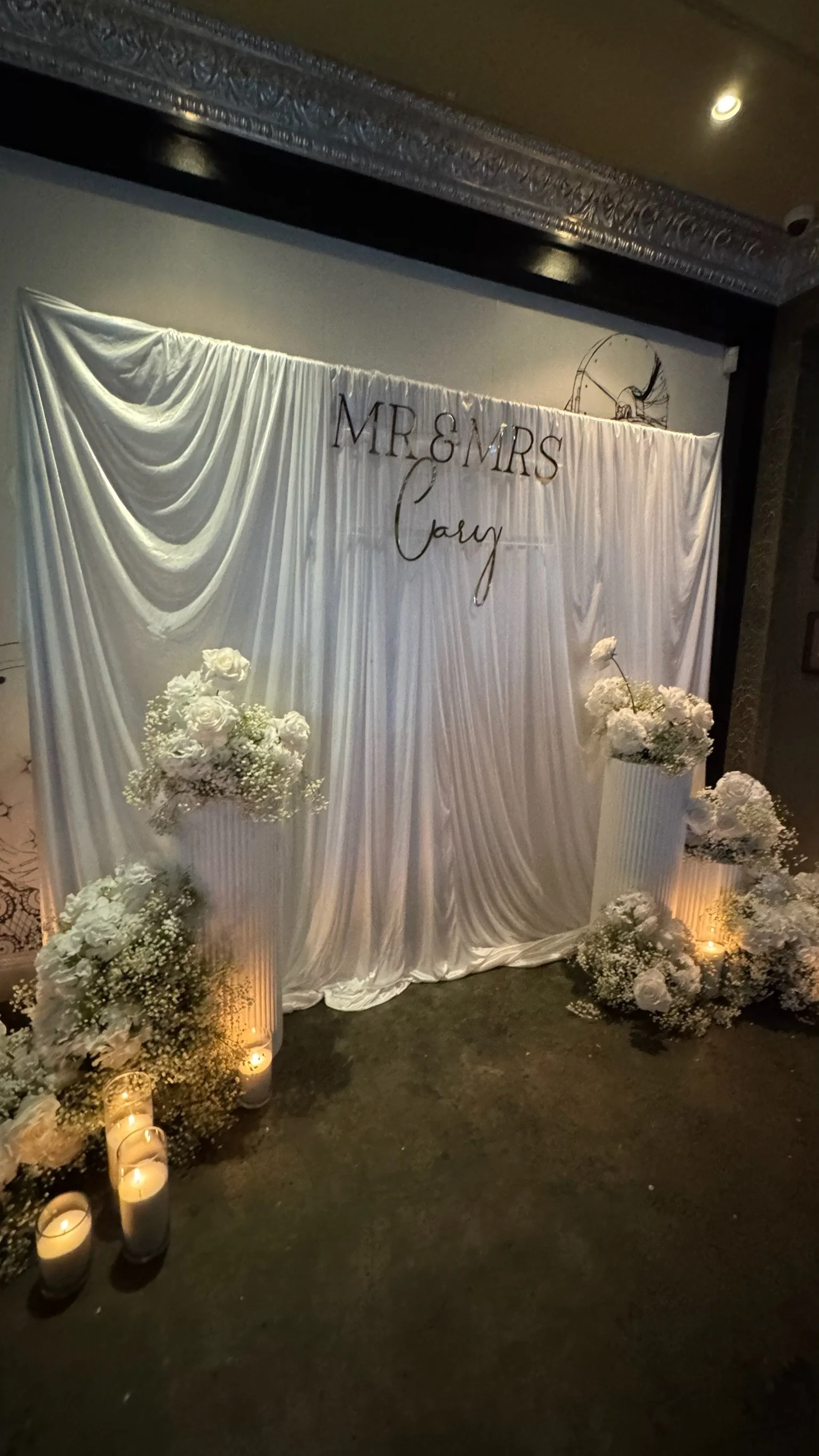 Wedding backdrop with white draped fabric, floral arrangements, candles, and the sign 'Mr & Mrs Cary' in elegant lettering.