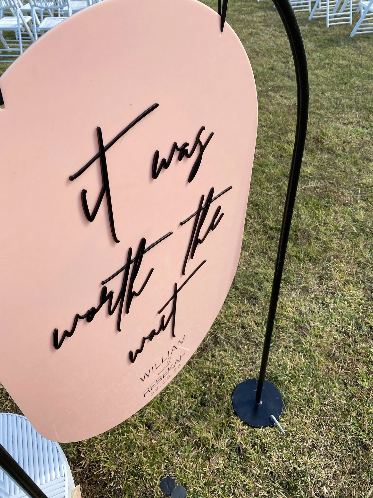 A pink sign with the handwritten phrase 'it's okay about the wait' in black cursive on a grassy outdoor area with white chairs in the background.