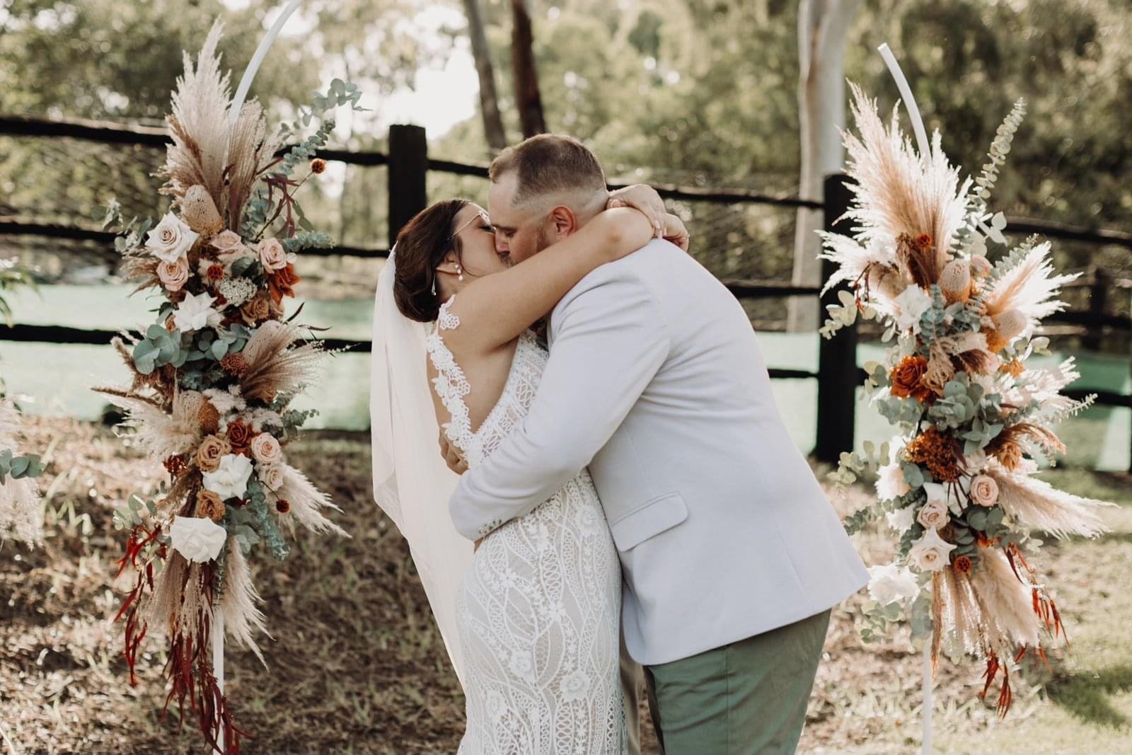 A bride and groom sharing a kiss during an outdoor wedding ceremony, standing between two decorated floral arches with pink, cream, and rust-colored flowers and greenery.