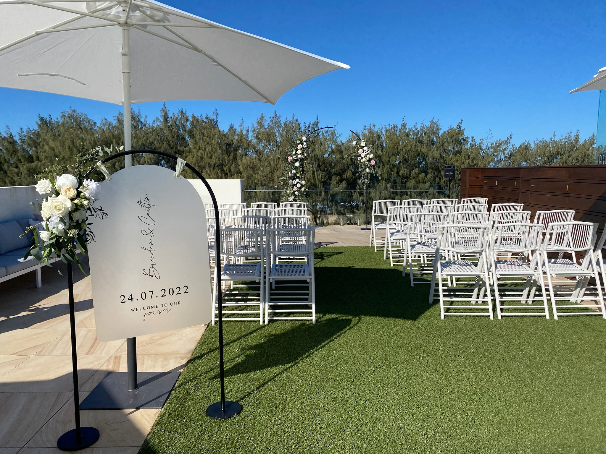 Wedding ceremony setup on a rooftop with white chairs arranged in rows, floral decorations, a welcome sign, and umbrellas under a clear blue sky.