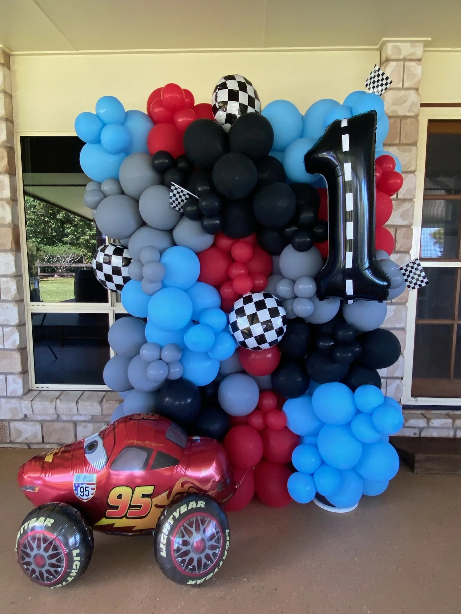 Balloon arrangement for a race car theme with red, black, blue, and gray balloons, a large number one balloon, checkered flags, and a Lightning McQueen race car balloon at the base.