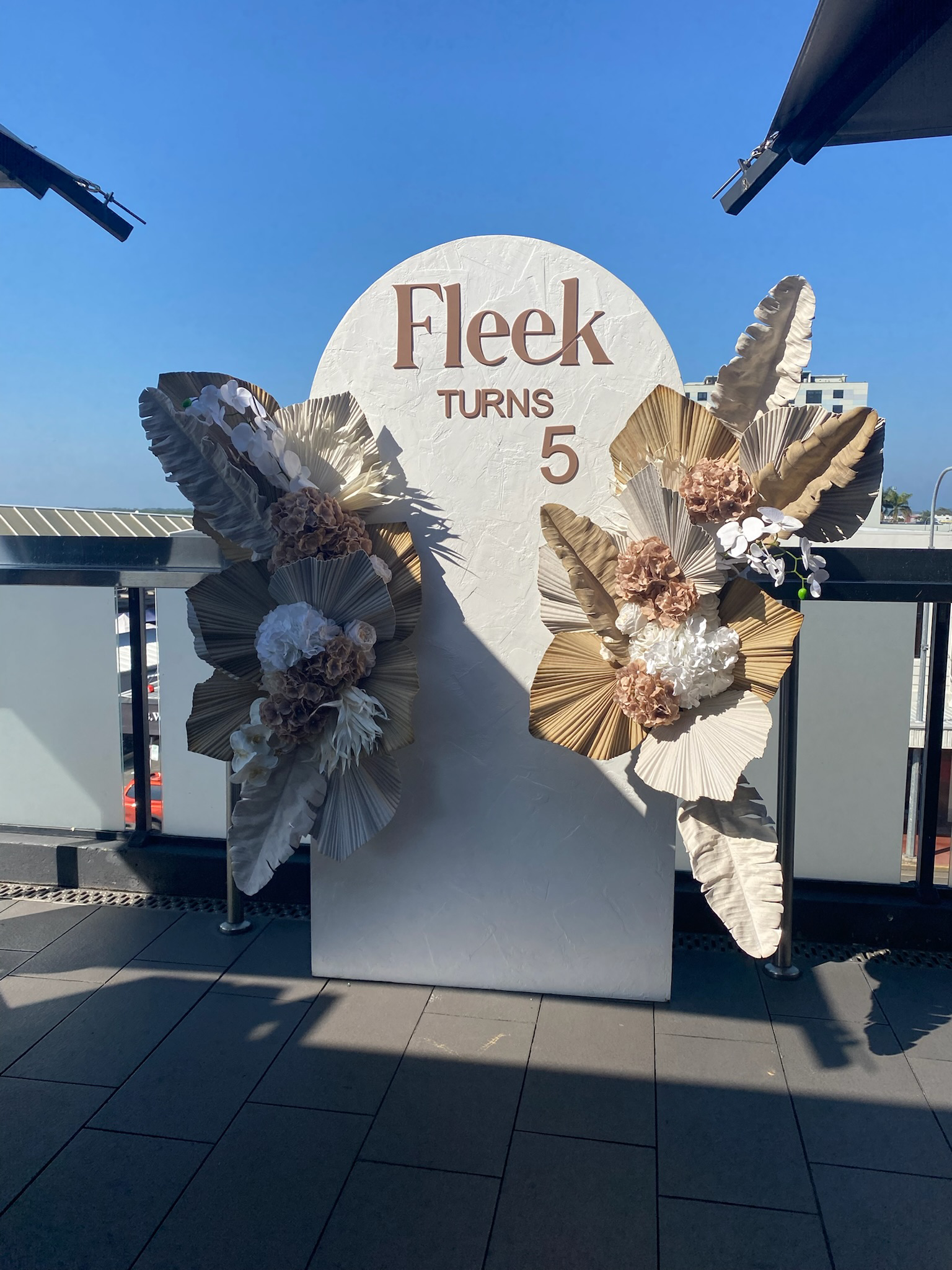A decorative sign with the text 'Fleek Turns 5' surrounded by beige, white, and brown paper flowers and leaves on an outdoor terrace with a blue sky in the background.