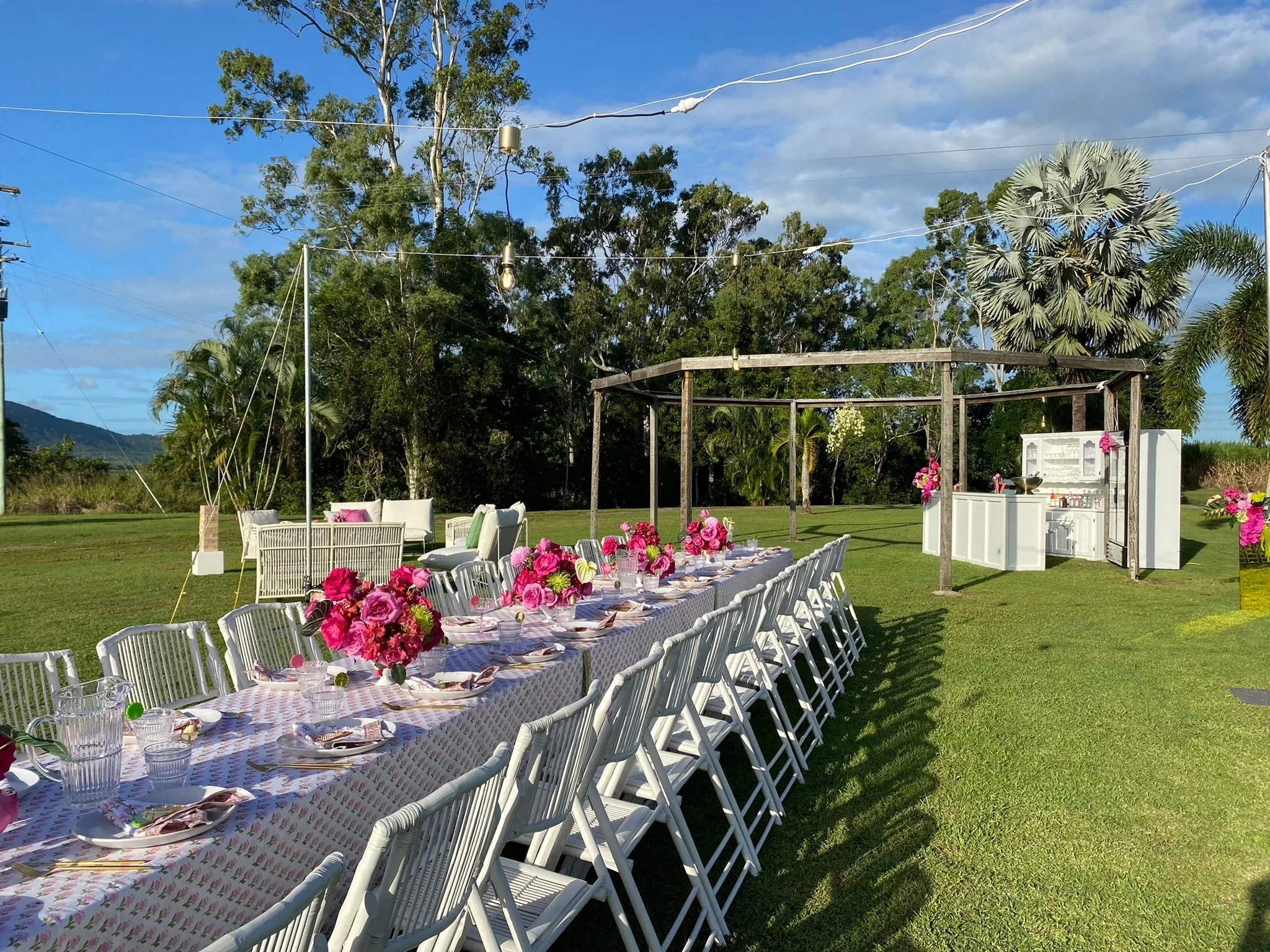Outdoor setting with a long table decorated with pink floral centerpieces, white chairs, and a white bar or food station in the background, surrounded by trees on a sunny day.