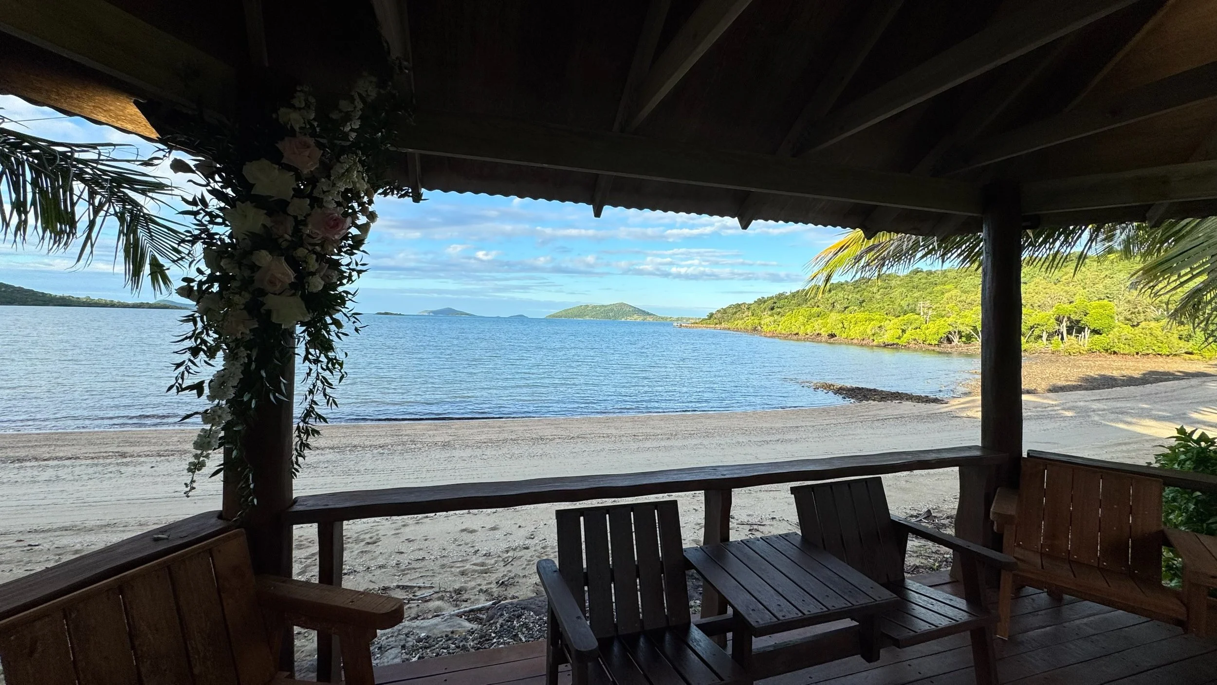 Beach view from shaded wooden hut with benches, decorated with flowers, overlooking calm water and islands in the distance.