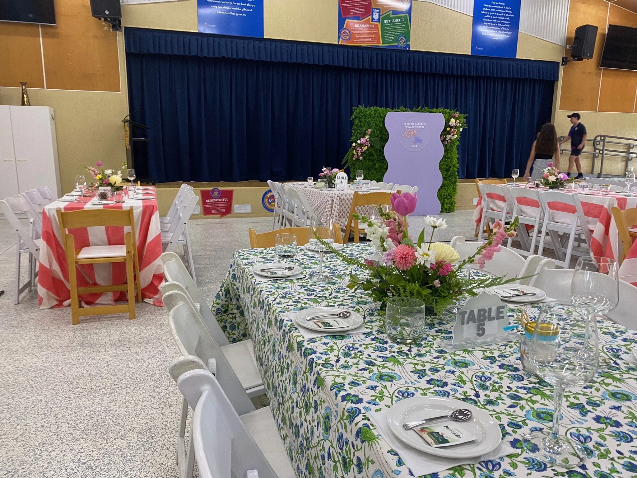 Event hall set up for a celebration with decorated tables, floral centerpieces, and a stage with a backdrop, preparing for a ceremony or banquet.