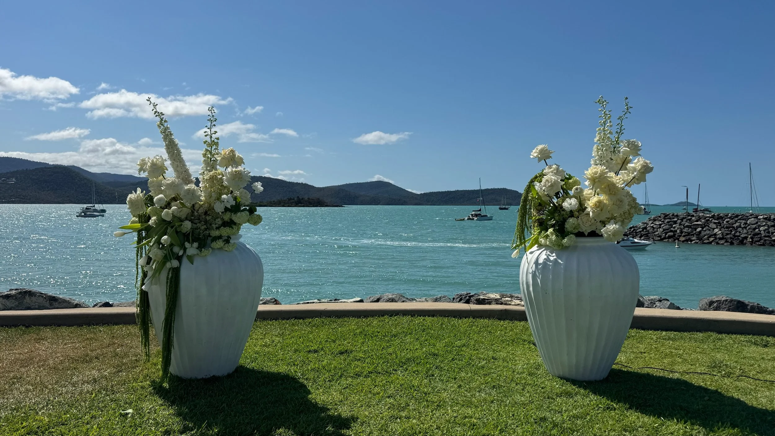 Two large white vases filled with white flowers and greenery, set on a grass lawn near a body of water with sailboats, on a sunny day with a blue sky and clouds.