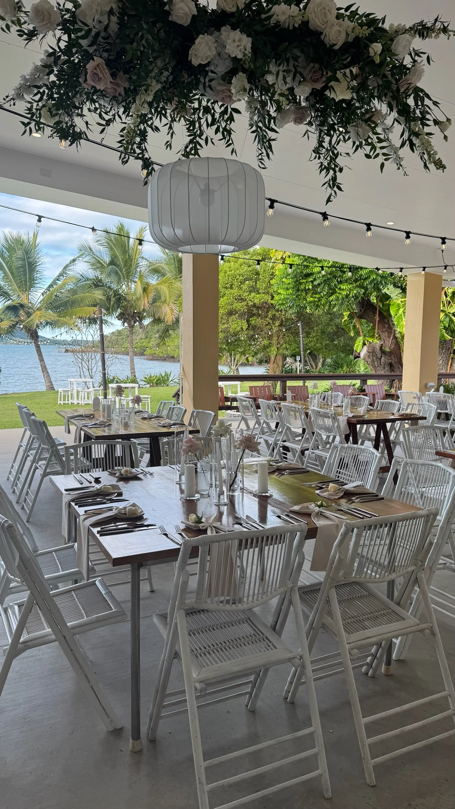 An outdoor restaurant setup near a lake with trees, white chairs, tables decorated with flowers and candles, and a large white lantern hanging from a floral arrangement ceiling.