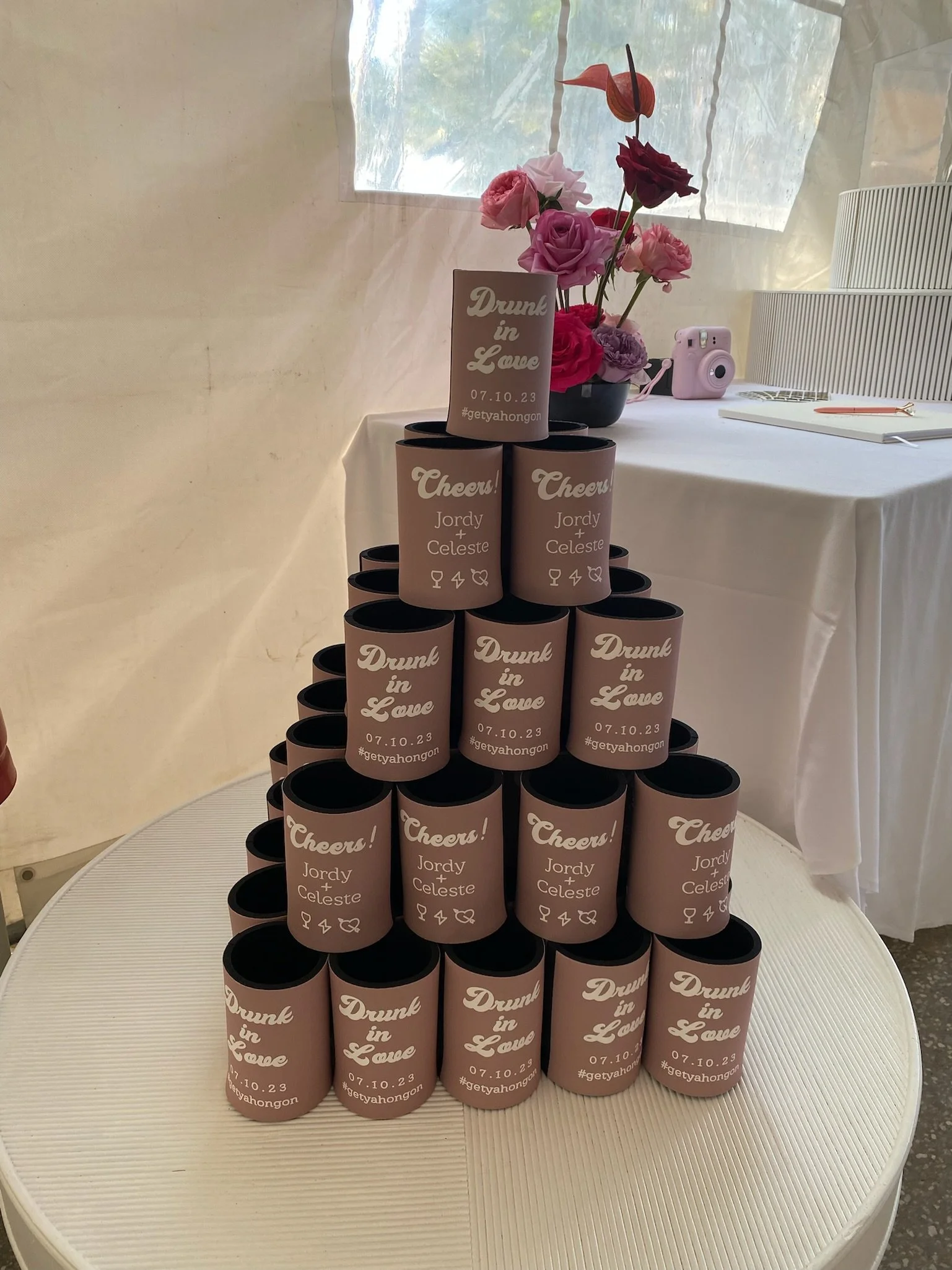 A pyramid of pink cans on a white table with a small sign on top reading 'Drunk in Love 07.10.23 #getyahongon.' Behind the pyramid is a vase with pink roses and an orange flower, a pink camera, and a notebook with a pen. A sheer curtain and a window 