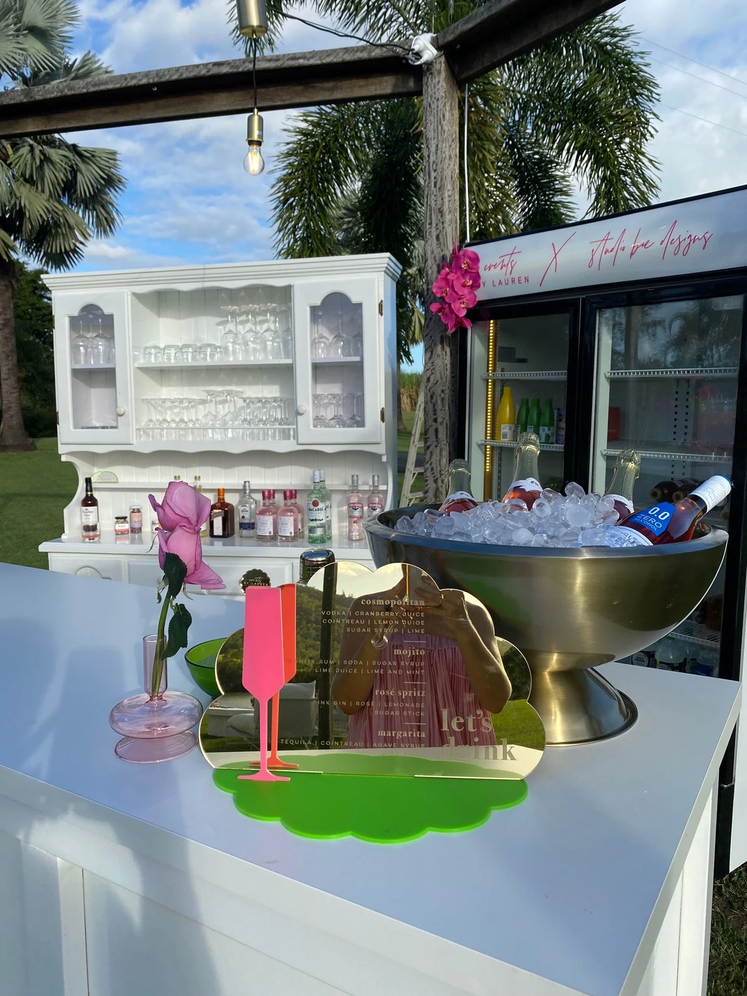 Outdoor bar setup with a white cabinet, drink bottles, a large bowl of bottled drinks on ice, a pink flower in a vase, and a colorful cocktail menu with decorative elements, under a wooden pergola with string lights, surrounded by palm trees.