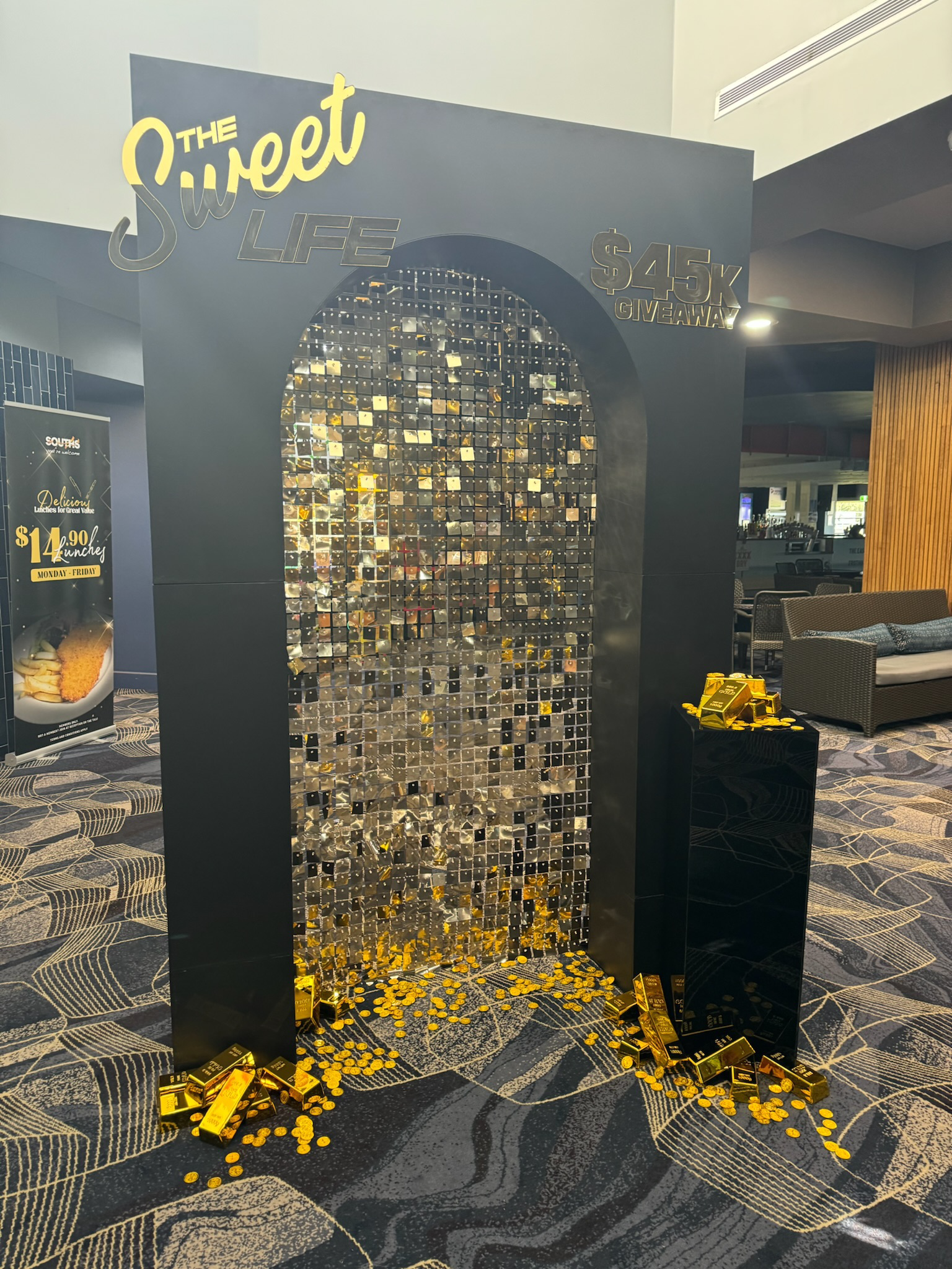 A promotional display with a black background and an arched mirror made of small mirror squares, surrounded by scattered gold bars and coins. Text reads 'The Sweet Life $45K Giveaway' at the top.