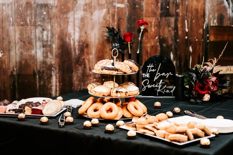 A table with assorted donuts, cookies, and chocolates against a wooden wall, decorated with flowers and a sign reading 'The Bar Sweet Kind'.