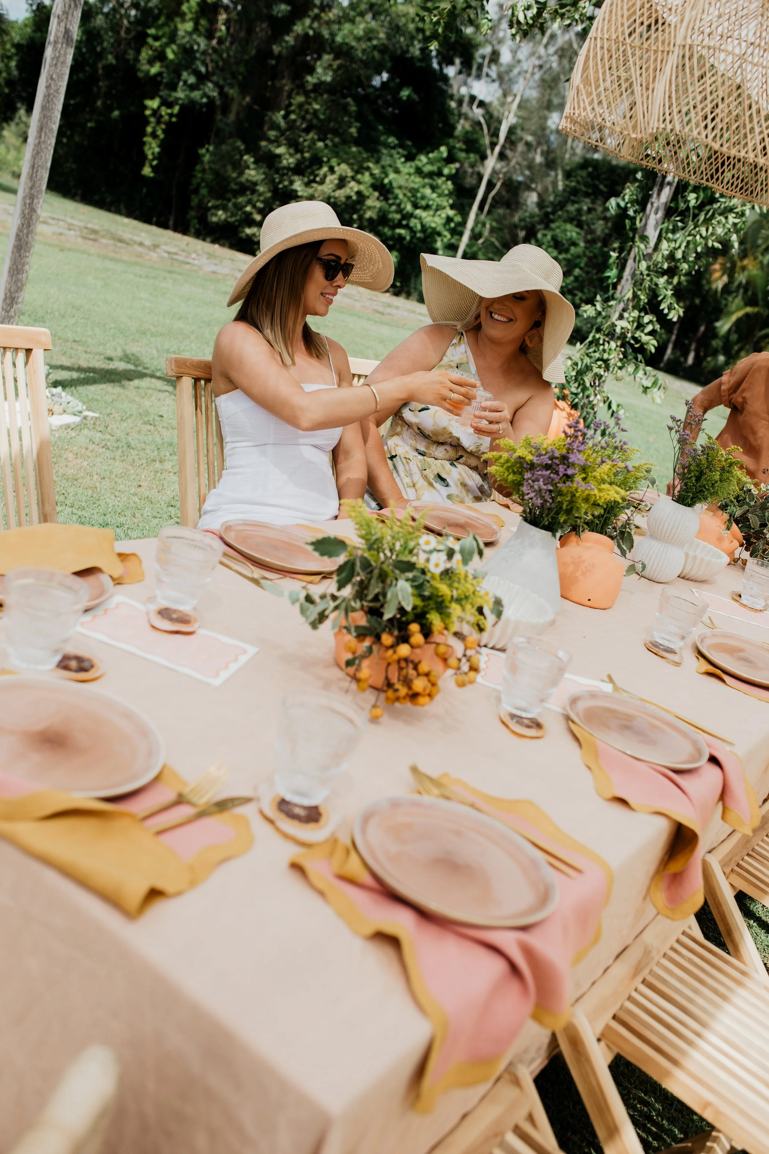 Two women sitting at an outdoor table, sharing a drink, with a decorated table setting including plates, glasses, and flowers, on a sunny day.