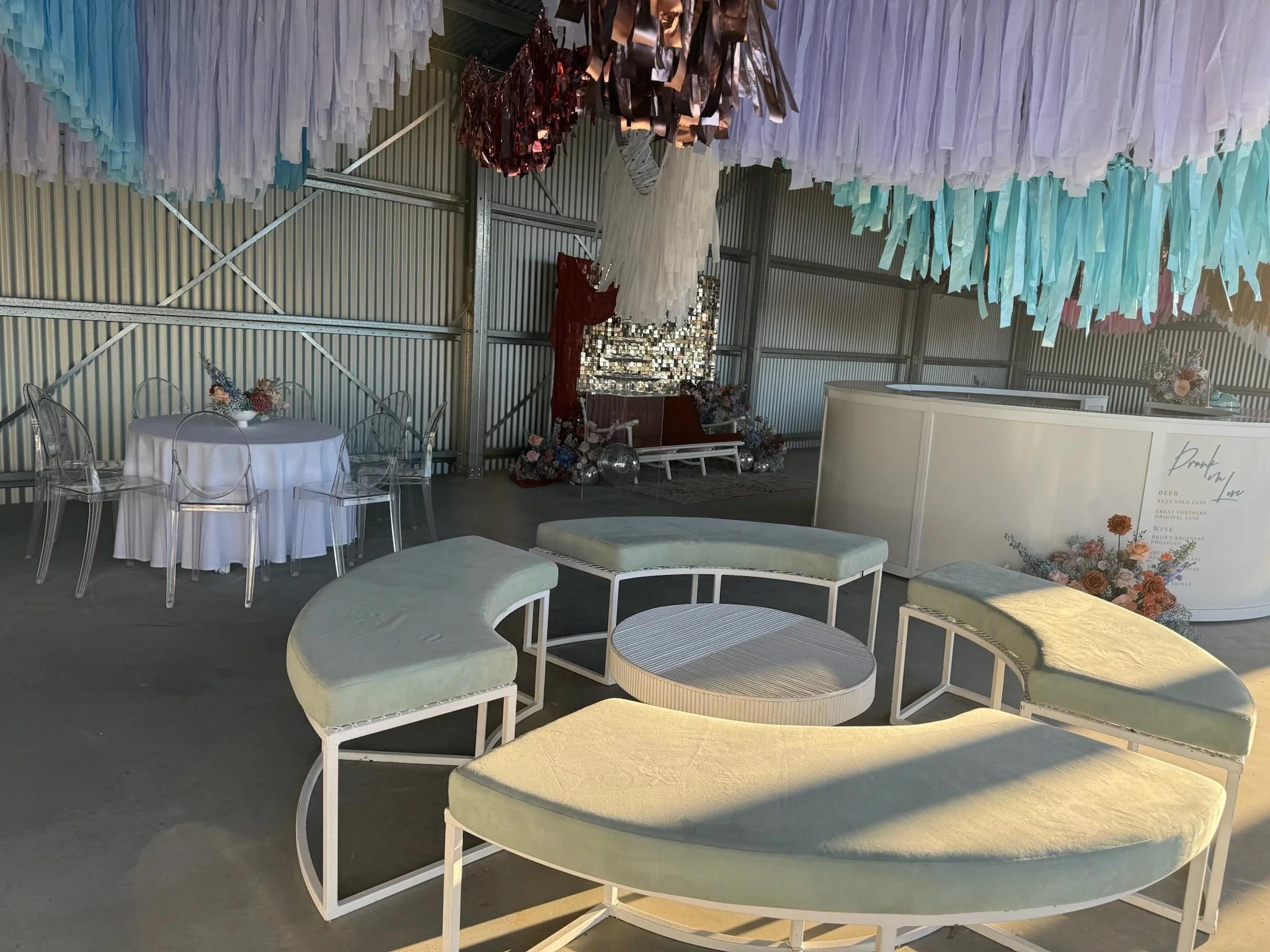 Indoor event space decorated with pastel-colored paper streamers hanging from the ceiling, offering a mix of seating options including a semi-circular sofa with pastel cushions, a round table with clear chairs, and a bar area with floral arrangements
