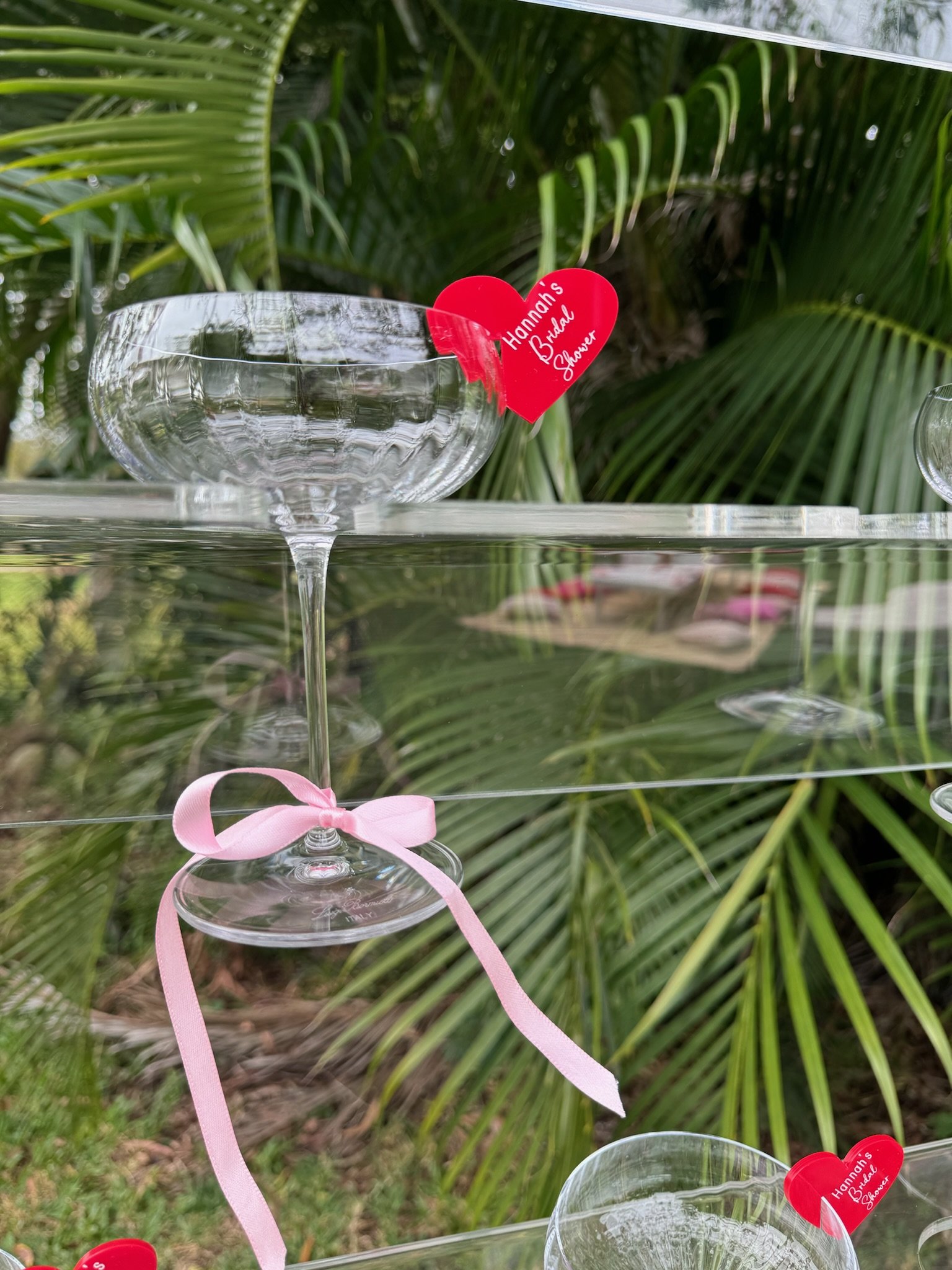 A clear glass bowl on a glass shelf with a pink ribbon tied around its stem, set outdoors against green palm leaves. There are red heart-shaped tags with white writing attached to the bowl, indicating it is a bridal shower gift from Hannah.