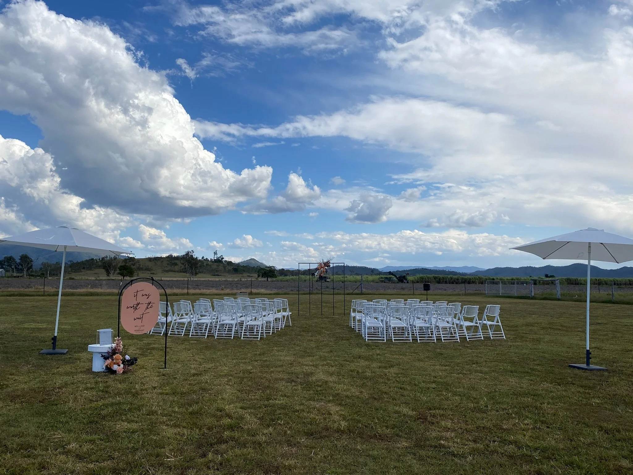 Outdoor wedding setup with white chairs arranged in rows on a grassy field, decorated with floral arrangements and umbrellas, under a partly cloudy sky with mountains in the background.