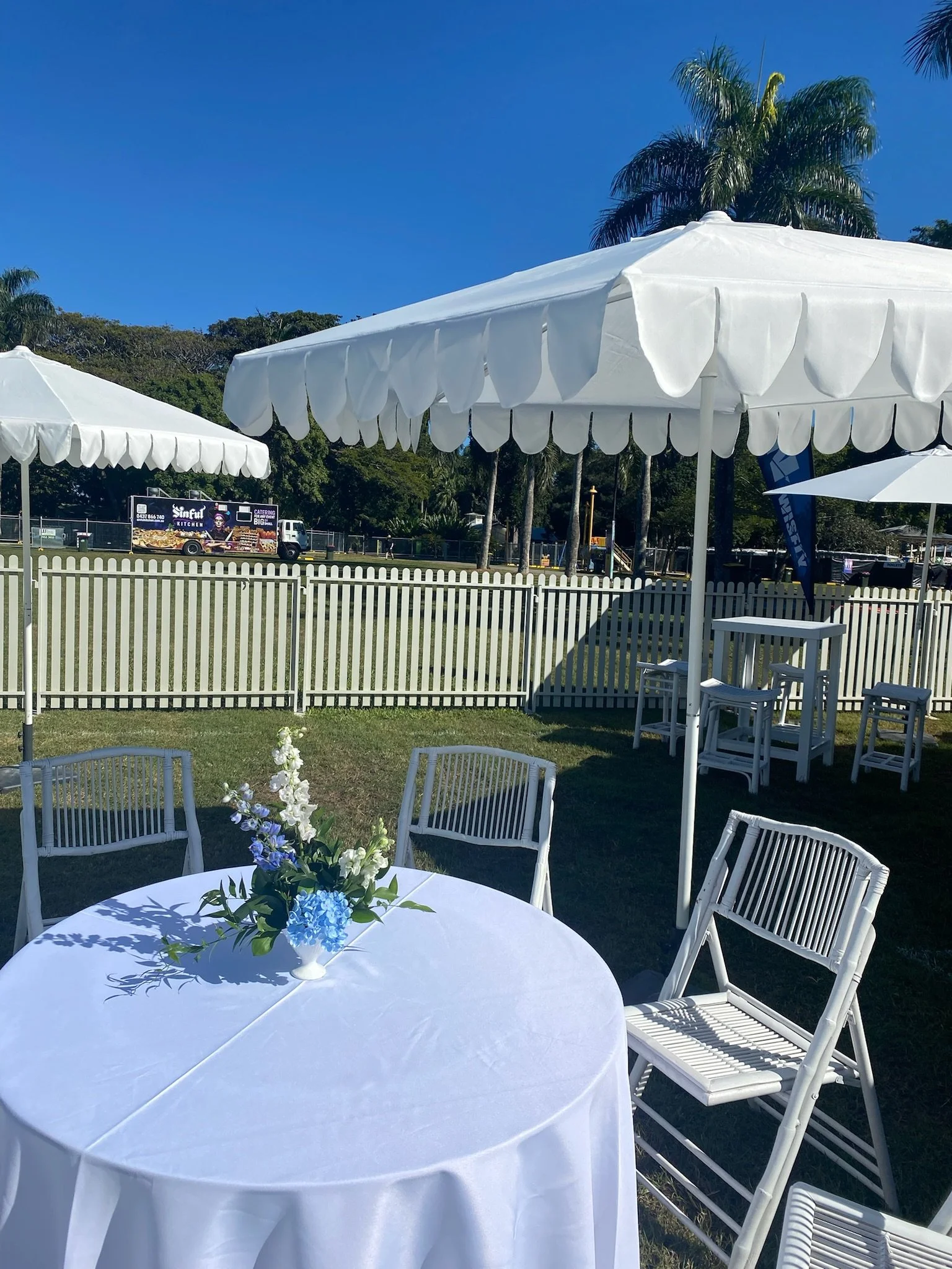 Outdoor event setup with white round table, white chairs, a flower centerpiece, white umbrellas, and a white picket fence against a backdrop of trees, blue sky, and palm trees.