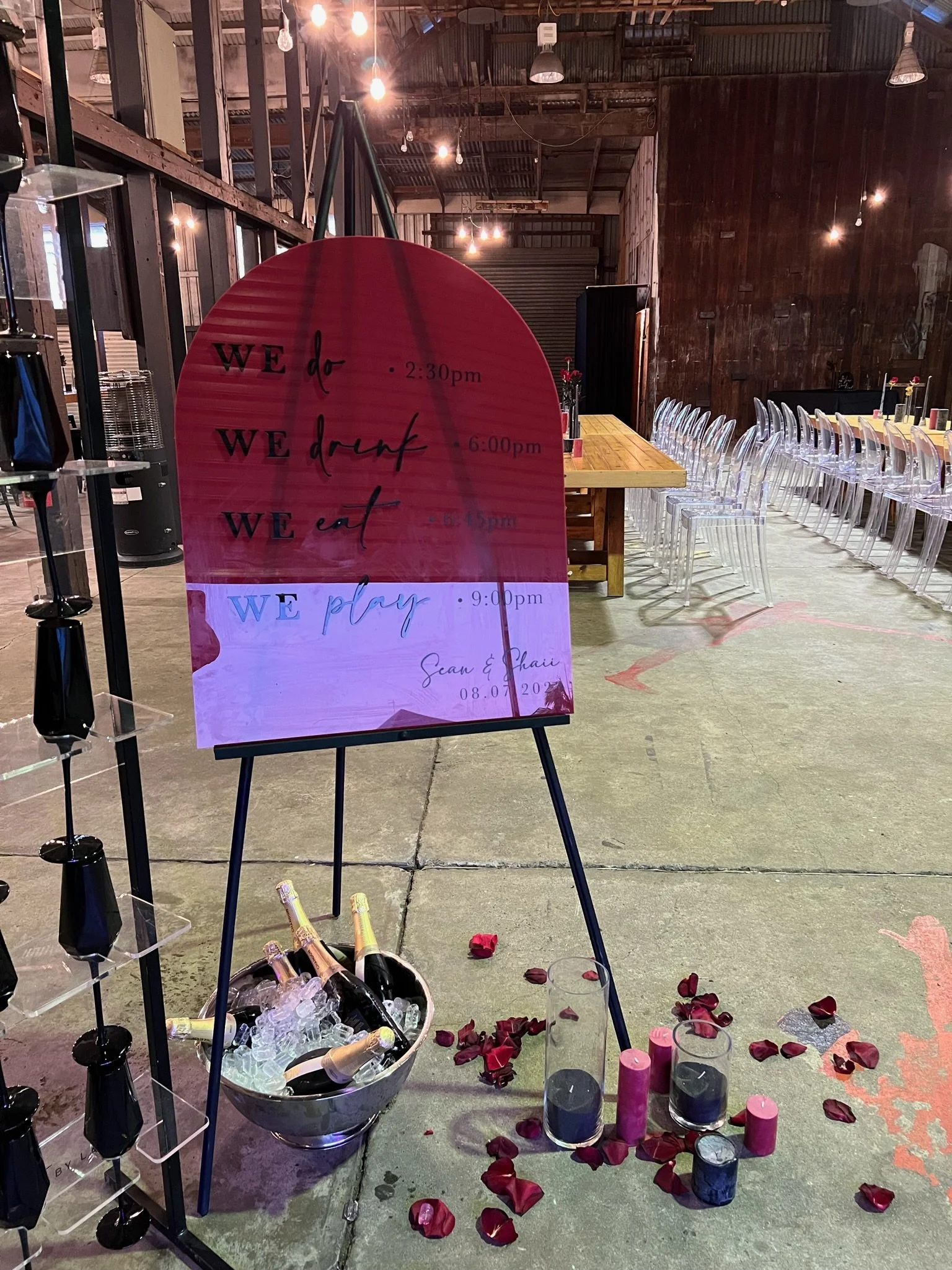 Event signboard outlining the schedule for the day with times and activities, surrounded by wine bottles in a bucket, scattered candles, and rose petals on the floor in a rustic indoor venue with string lights and transparent chairs.